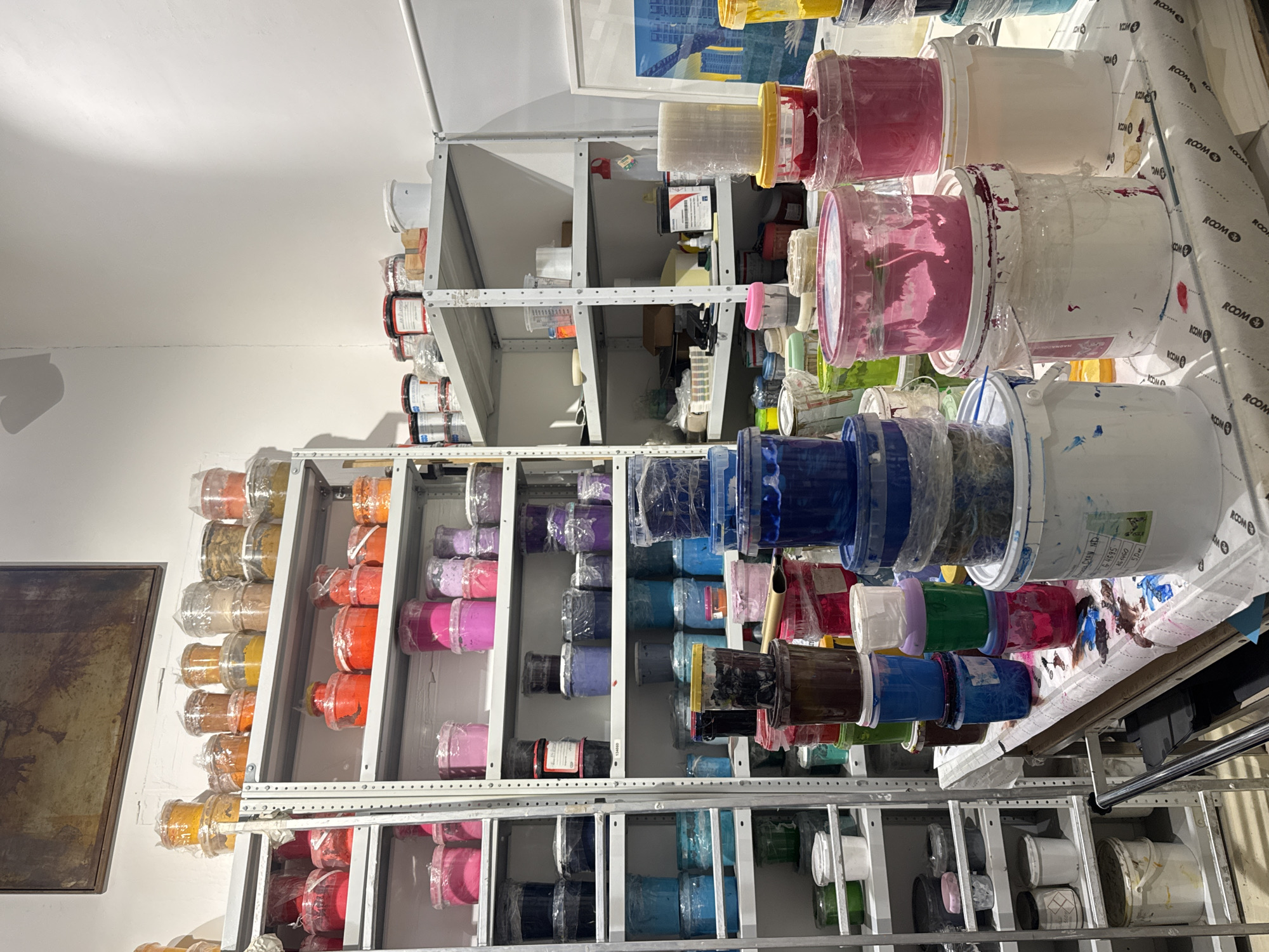 The image depicts a storage room filled with shelves holding various sizes of buckets filled with what appears to be paint or ink in a spectrum of colors. The buckets are mostly sealed with plastic wrap, and many have drips and splatters on the exteriors. The shelves are made of metal, likely steel or aluminum, and are adjustable. The room seems to be a workshop or studio, perhaps belonging to an artist or designer. There is a framed artwork or photograph hanging on the wall to the left. The exact location in the city of Unknown, Unknown, is not ascertainable from the image.