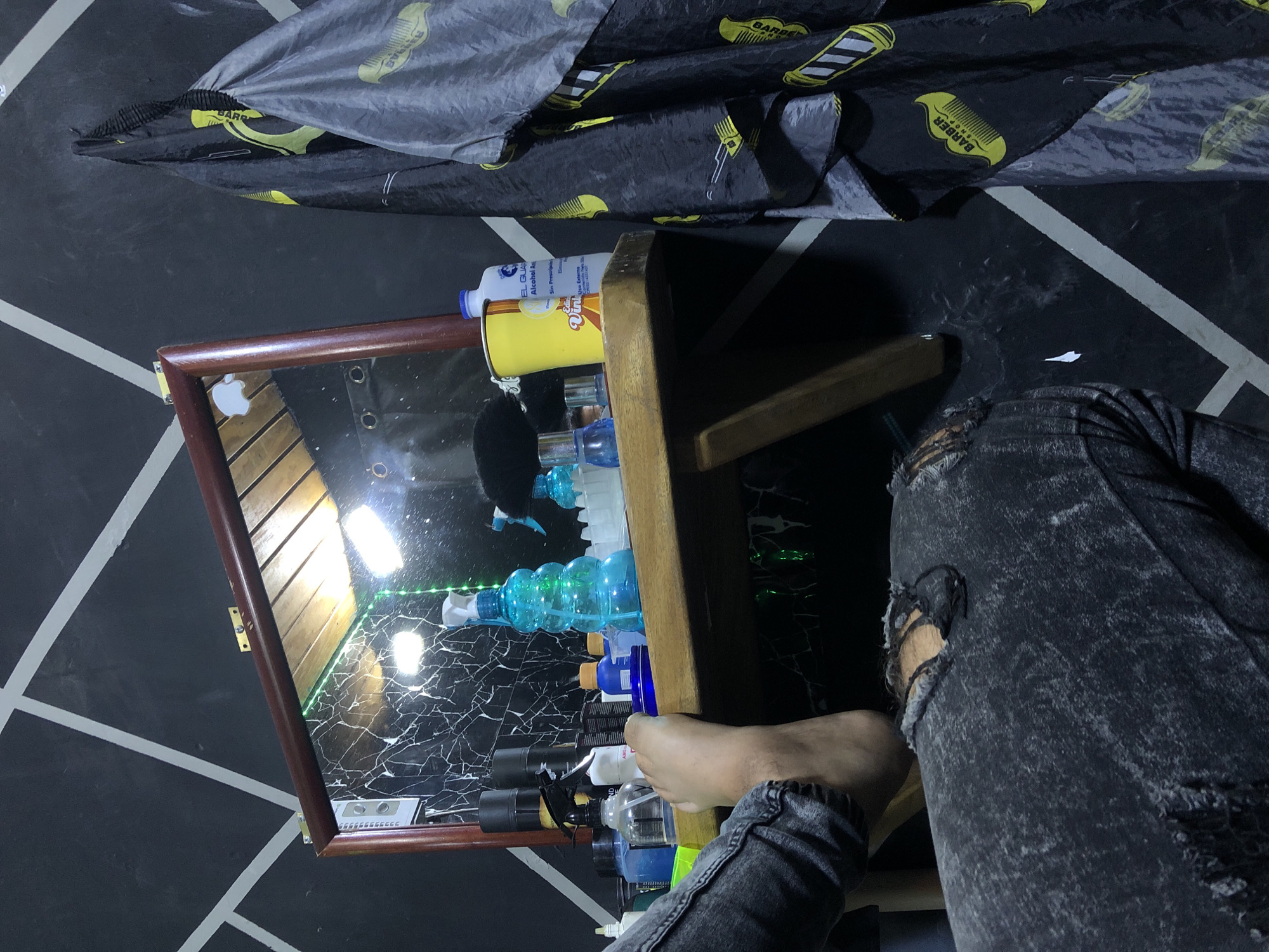 The image shows a small wooden table with a mirrored surface, cluttered with various bottles and containers. The table is placed on a floor with a black and white geometric pattern. A person's leg, clad in ripped jeans, is visible near the table. A barber's cape, patterned with combs, is draped in the background. The location is unknown, there is no way to confirm it is in Altagracia, Venezuela.