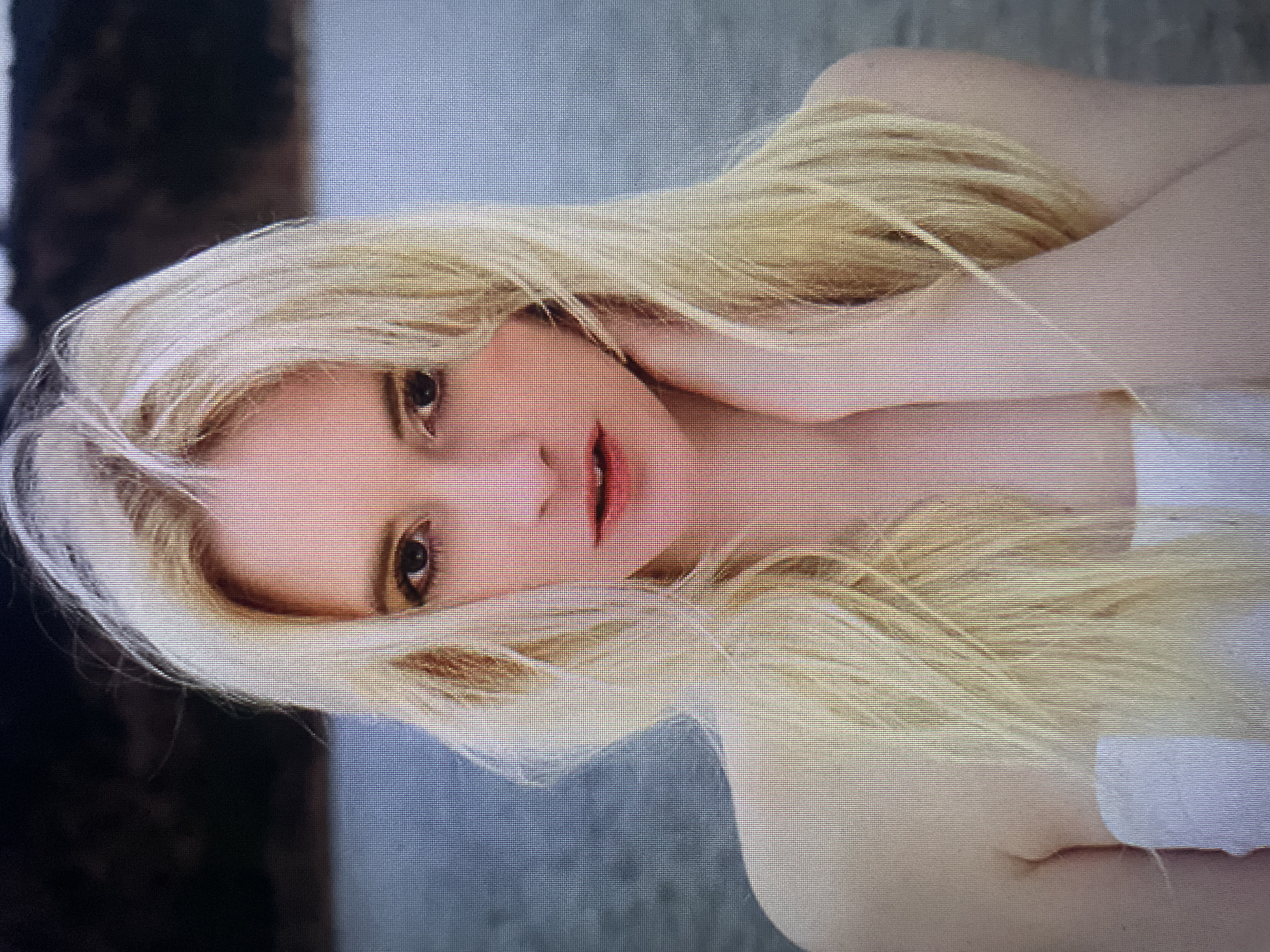 The image is a close-up, head-and-shoulders portrait of a young Caucasian woman with long, blonde hair. She is looking directly at the camera with a neutral expression. Her skin is fair, and she is wearing light makeup, with a subtle sheen on her eyelids and reddish lipstick. She has a white garment or wrap around her upper chest and shoulders. The background consists of a body of water with a darker, blurred area behind it, likely representing land or vegetation. The image quality is somewhat grainy, suggesting it might be a photograph taken from a screen or a low-resolution source. There is no indication of the specific location in Kołobrzeg, Poland.