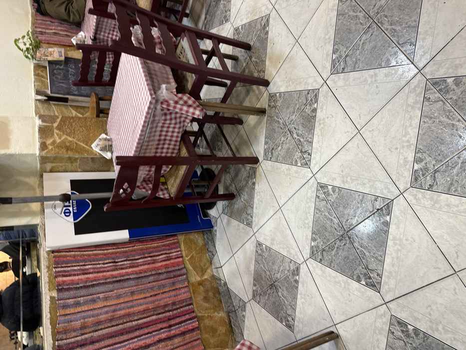 This media file captures the interior of what appears to be a casual restaurant or dining area, likely in Dafni, Greece, taken from a slightly elevated and angled perspective.
The scene features several dining tables, each adorned with a red and white checkered tablecloth. Matching wooden chairs with carved backrests are arranged around the tables. On one table in the upper left, a small green potted plant sits, and paper napkins are visible on multiple tabletops. A person is partially visible sitting at a table in the upper left.
The floor is covered with a distinctive geometric pattern of light and dark grey tiles, forming a repeating star-like or diamond motif. Along the left side of the image, the lower portion of the wall is clad with light brown, textured stone. A vibrant, horizontally striped rug in various shades of red, purple, and brown is laid on the floor adjacent to the stone wall.
To the left, a large white and blue object, possibly a display cooler or sign, stands against the wall. Text on this object clearly reads "ΦΩΣΣΙΕ" (FOSSIE) and "ESSIE", indicating a brand. Another person, wearing a dark jacket, is partially visible near this object. A dark menu board, leaning against the stone wall, shows some handwritten text, including "3,00€", suggesting prices. The overall ambiance is rustic and inviting.