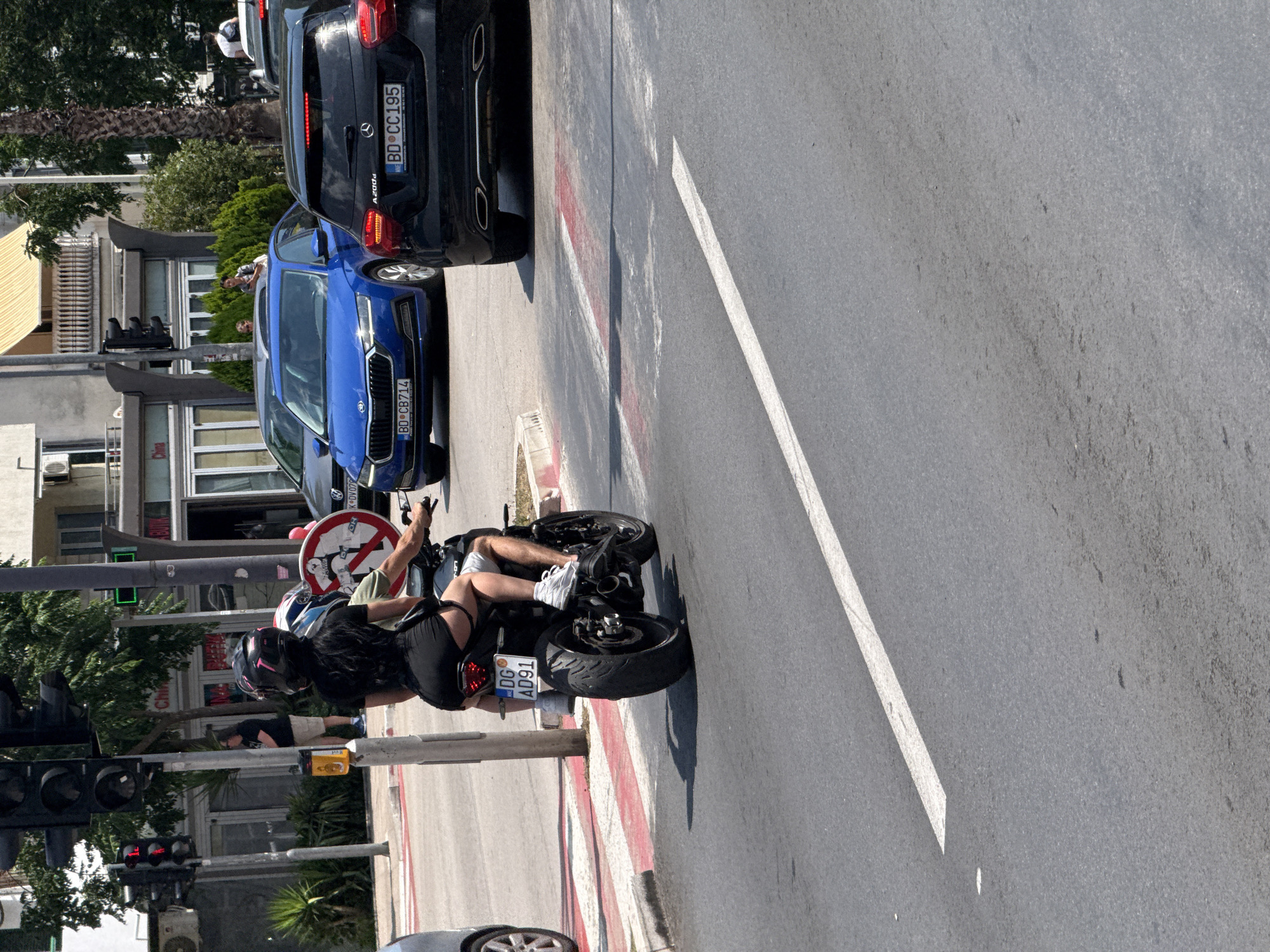 In Budva, Montenegro, two individuals, a driver and a passenger, are riding a black motorcycle on a city street. The driver, wearing a helmet with pink accents and a black shirt and shorts, is operating the motorcycle. The passenger, also wearing a helmet, is seated behind the driver. The motorcycle has a license plate that reads "DG AD91". The scene takes place at a street intersection with traffic lights, pedestrian crossing markings, and other vehicles in the background, including a blue Skoda and a black Mercedes. The surrounding environment includes buildings, trees, and a clear sky.
