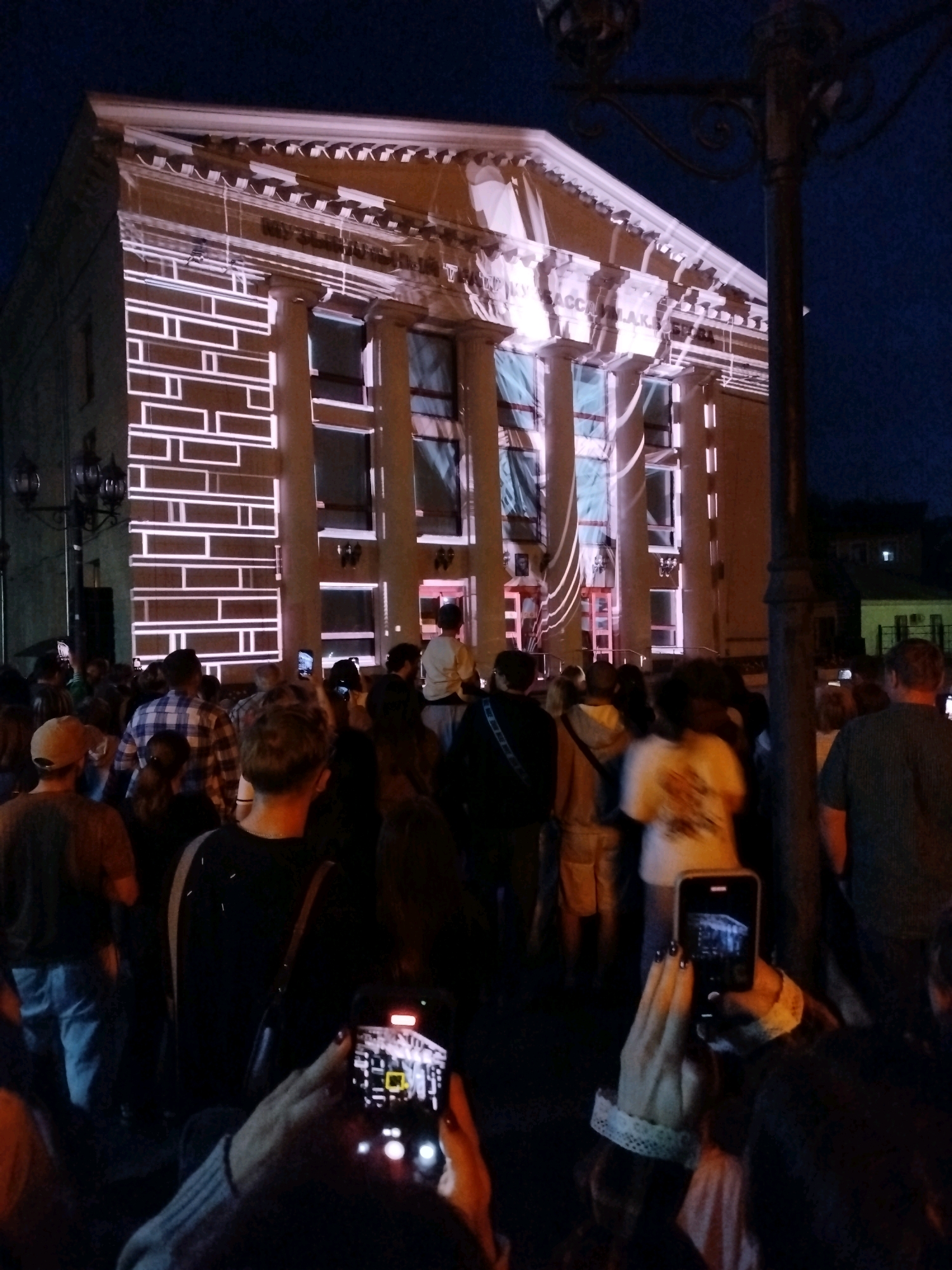 The image depicts a night scene in front of a building illuminated with a light projection. A crowd of people is gathered, many holding up their phones to capture the spectacle. The building appears to be a cultural institution, with columns and architectural details visible. The light projection on the facade creates a geometric pattern of white lines against a darker background. The sky is dark, suggesting it is nighttime. The overall atmosphere is that of an outdoor event or exhibition. The building in the image is identified as the Kemerovo State Philharmonic, located in Kemerovo, Russia.