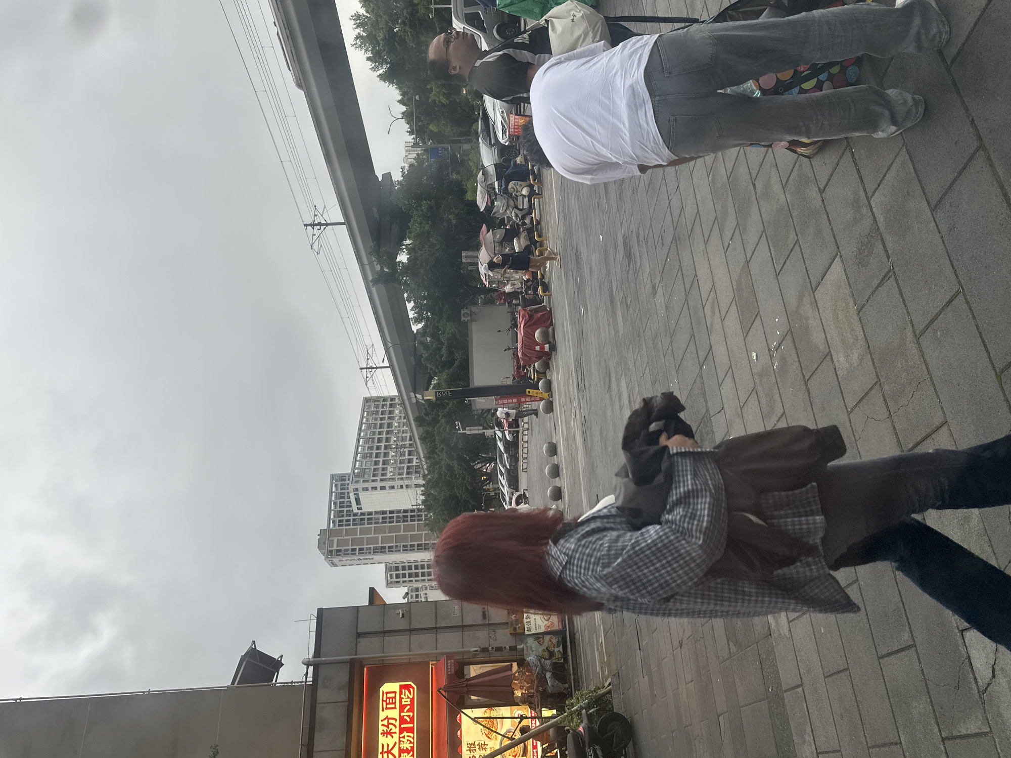 The image depicts an outdoor scene in what appears to be a city. In the foreground, a person with long, reddish-brown hair is viewed from behind, wearing a plaid shirt and holding a dark jacket. To the left and further back, another person is seated on the ground, wearing glasses and a dark t-shirt. Beyond them, a line of parked scooters is visible, some covered with umbrellas. In the middle ground, there is a street with vehicles and a row of buildings in the background, some of which are tall, modern structures. An elevated road or bridge structure runs across the upper portion of the image. The sky is overcast, suggesting a cloudy or possibly rainy day. The scene appears to be a public space, likely a sidewalk or street, with commercial establishments indicated by signage. Visible text includes Chinese characters on a red sign, which reads "天粉小面" (Tian Fen Xiao Mian). The overall atmosphere is mundane and urban, with no strong indicators of a specific event. The time of day is not clearly discernible but appears to be daytime.