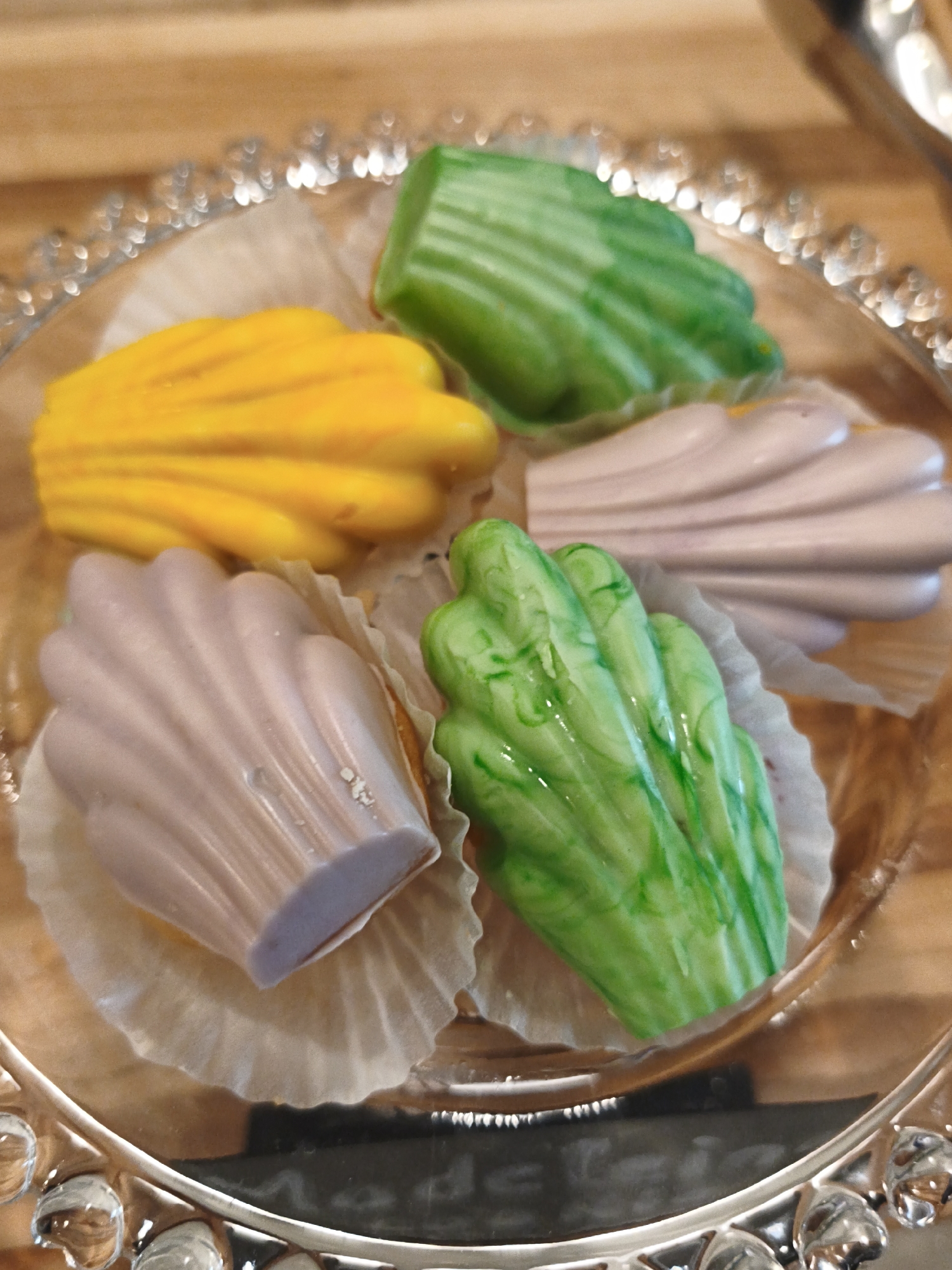 This is a close-up, overhead shot of four individually wrapped chocolates on a decorative, ornate silver platter. The platter has a raised, scalloped edge with small, circular embellishments. The chocolates are shaped like seashells or fans and are in various colors: yellow, pale purple, and two shades of green. The chocolates are placed in fluted paper cups. The background appears to be a wooden surface, possibly a table. There are no people or visible signs indicating a specific location in Bac Nha Trang Ward, Vietnam.