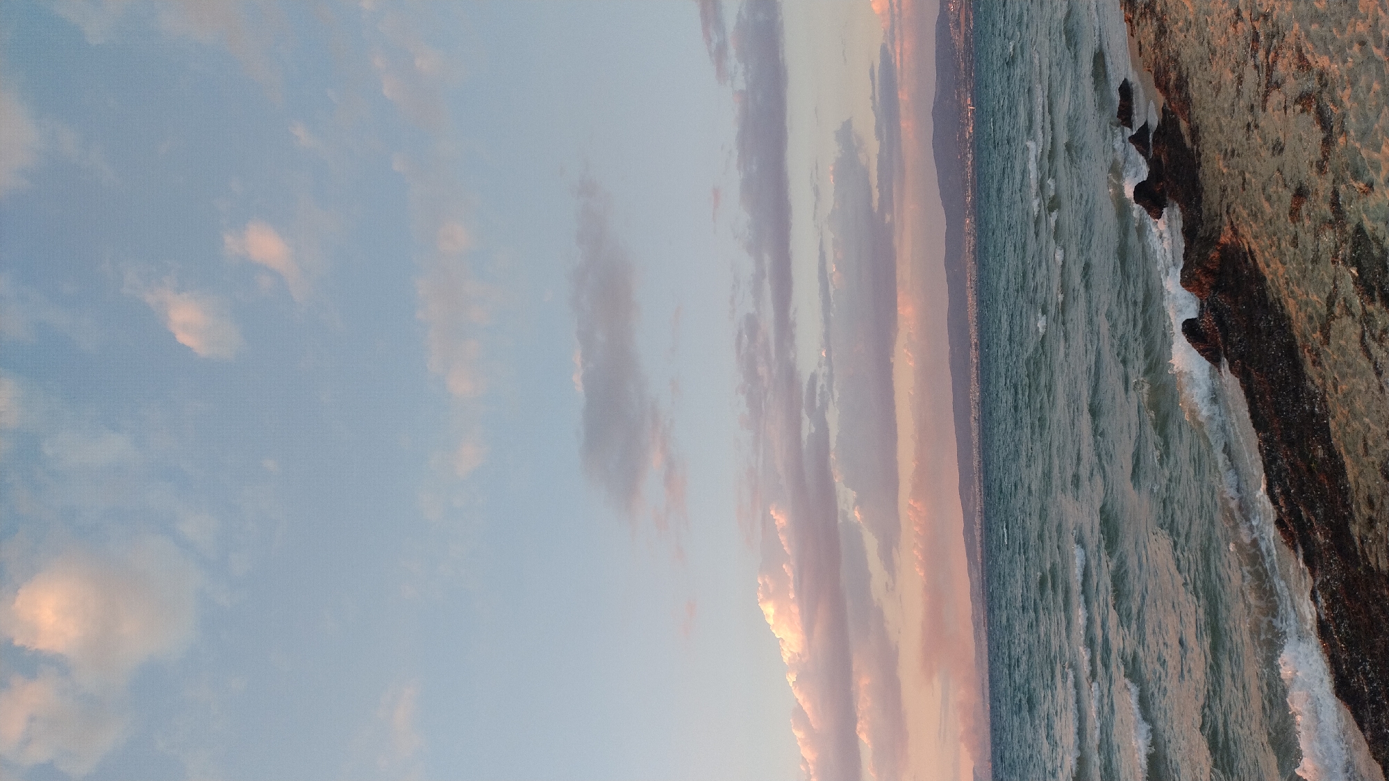 The image depicts a serene coastal scene at sunset in Neoi Epivates, Greece. The sky is a gradient of pale blue with scattered clouds tinged with pink and orange hues from the setting sun. The sea, a deep teal, has gentle waves lapping at the shore. The beach is composed of light brown sand, with some dark rocks and seaweed scattered along the waterline. In the distance, a coastline is visible, featuring a city with buildings and hills. The overall atmosphere is calm and tranquil, characteristic of a peaceful evening by the sea.