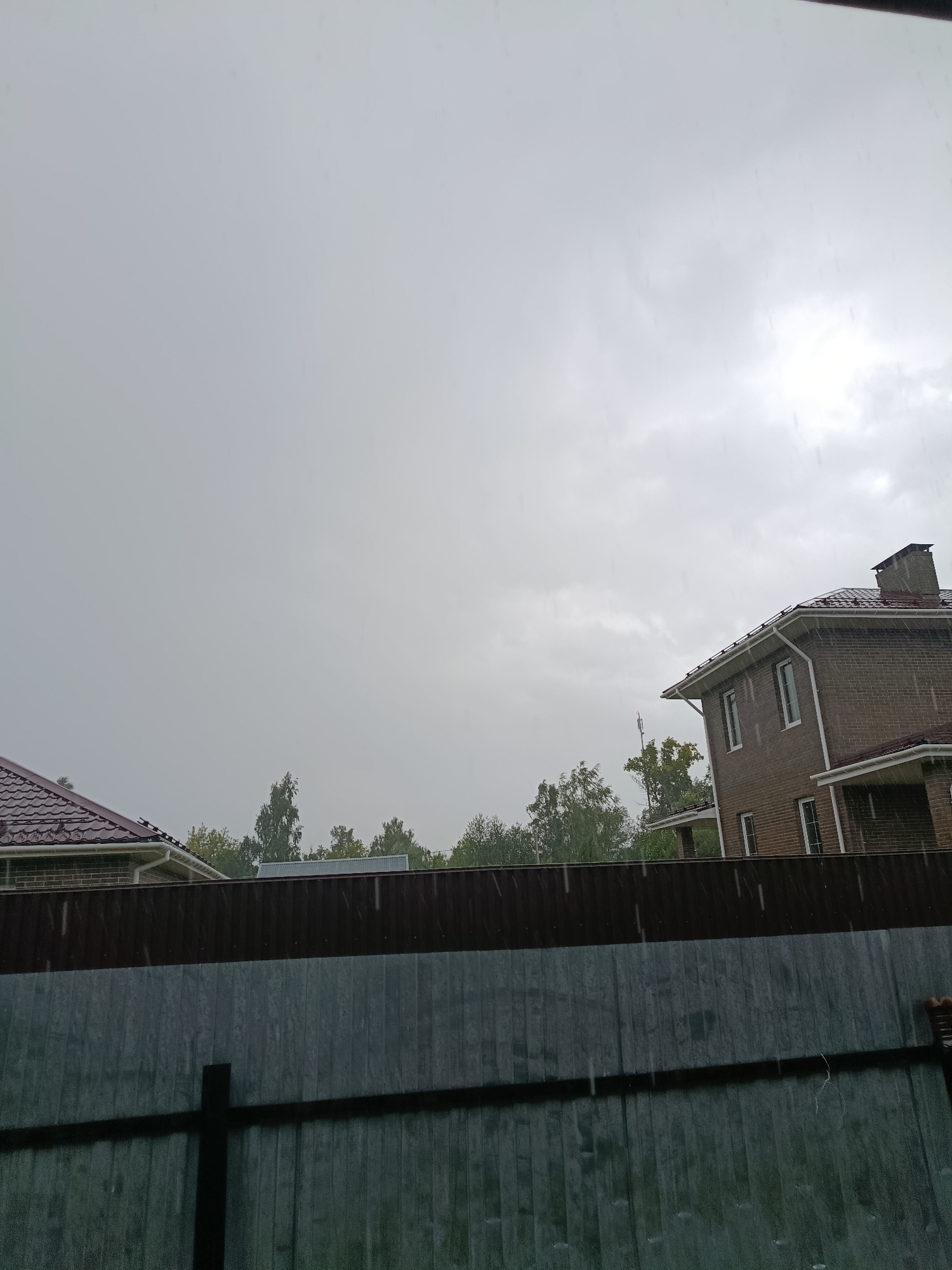 This image depicts a view of houses and a fence during a rain shower in the city of Vereya, Russia. In the foreground, a dark gray metal fence runs horizontally across the frame, with vertical supports visible. Behind the fence, a darker brown corrugated metal fence is situated. Beyond the fences, trees with green foliage are sparsely distributed against a bright, overcast sky.
On the right side of the image, a two-story house constructed of brown brick with a dark brown tiled roof is visible. The house has white window frames. To the left of this house, another house with a dark brown tiled roof and white trim is partially visible, along with a portion of a brick wall. A cell tower with a red antenna is located behind the trees to the left of the brick house. The sky is predominantly gray, with patches of lighter gray suggesting the presence of clouds. Visible rain streaks are apparent throughout the image, indicating active precipitation. The overall scene is one of a residential area experiencing inclement weather.