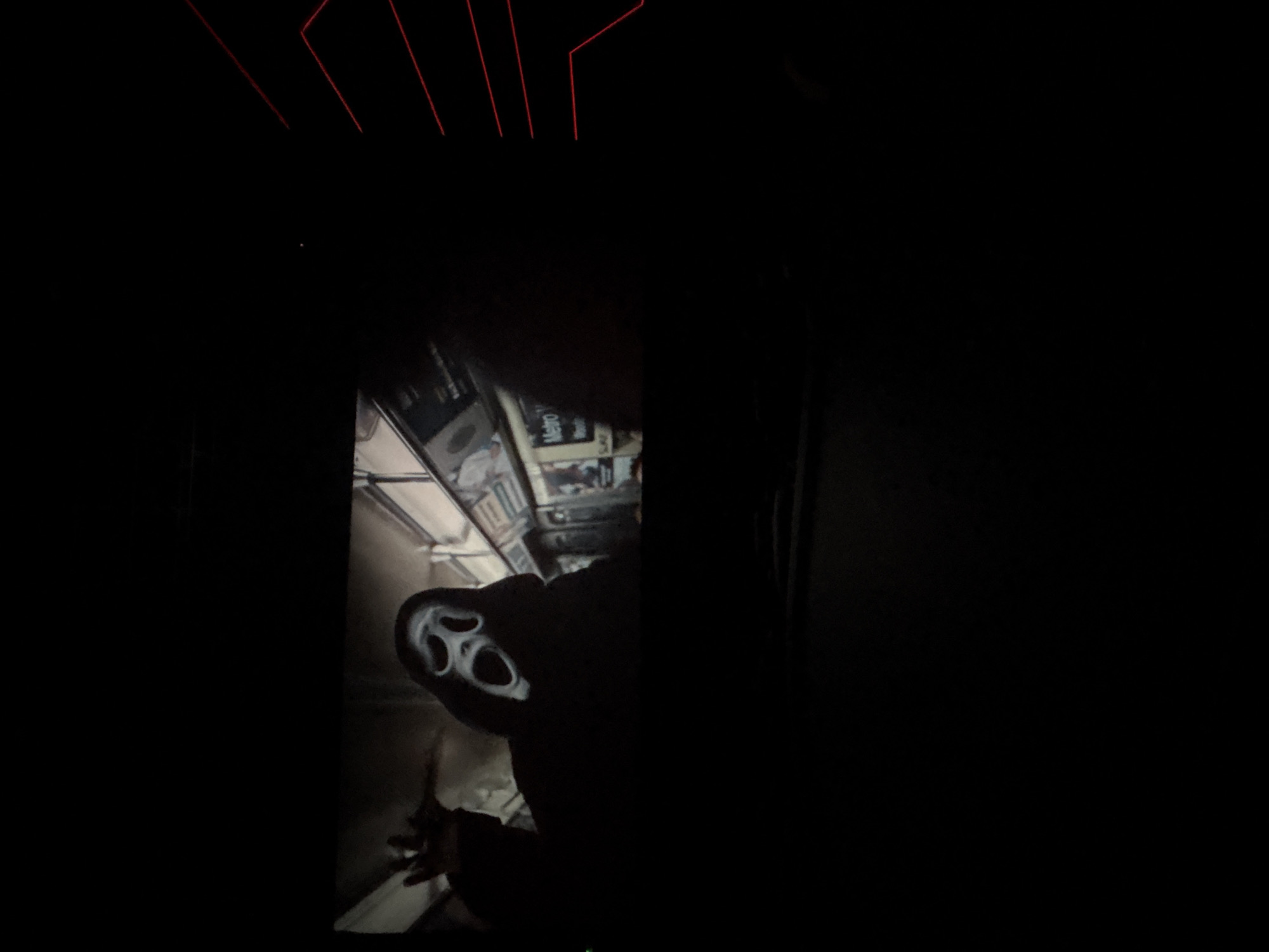 The image shows a person wearing a "Ghostface" mask, likely taken in a dark or dimly lit environment. The person is wearing dark clothing, obscuring most of the body. The setting includes a structure with a sign that is slightly visible and indicates a subway or Metro station. The background is mostly dark, though there are hints of possible interior elements. There are also angled red light fixtures near the top of the image. The information available does not allow me to precisely identify the location as Varna, Bulgaria.