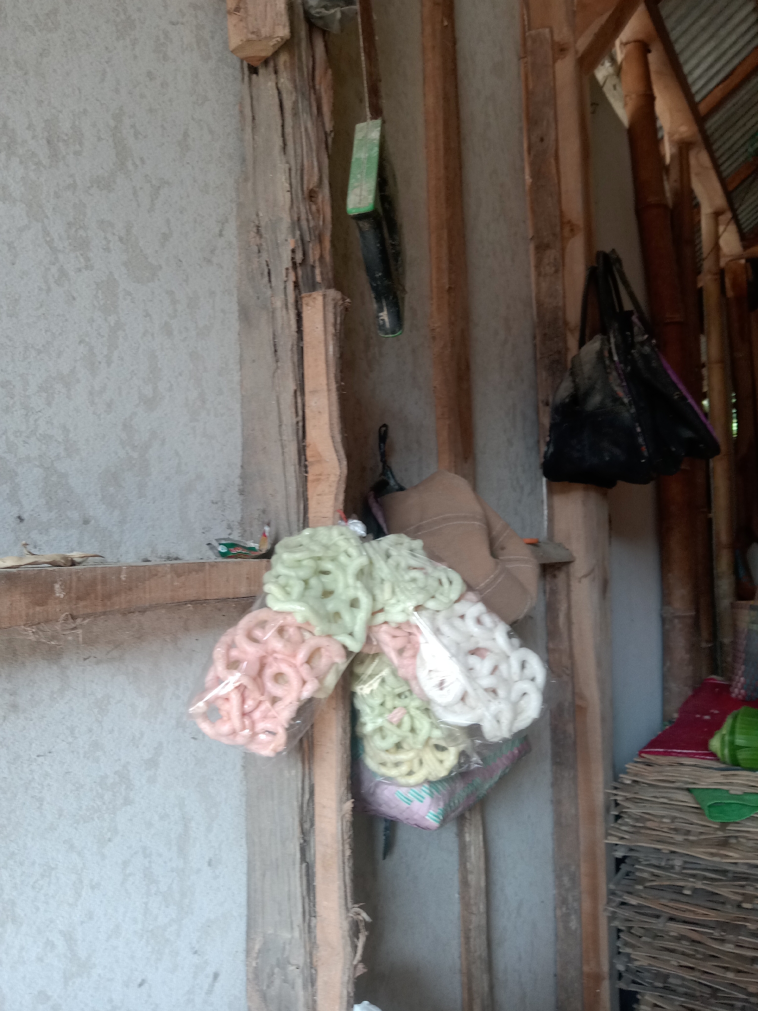 The image captures a cluttered, rustic interior scene, likely in a rural or semi-rural dwelling in Pacing, Indonesia. In the foreground, a pile of colorful crackers, possibly kerupuk, are packaged in plastic and arranged in plastic bags. They are predominantly pink and light green, with some white ones visible. These are stacked on top of a patterned fabric.
The background reveals a rough, unfinished wooden structure, with exposed beams and posts. One of the wooden posts runs vertically down the center of the frame. To the left, a light gray, textured wall is visible. Above the crackers, a brown textile, possibly a bag or fabric, is draped over a wooden beam. Further back and to the right, a black bag hangs from a wooden support. The structure extends into the background where vertical bamboo poles are visible, suggesting a building with a natural or partially constructed material. A corrugated metal roof is partially visible at the top right, hinting at an outdoor element or a partially covered area. The overall impression is one of a storage or utility space, with everyday items stored in a somewhat unorganized manner. There are no people present in the image.