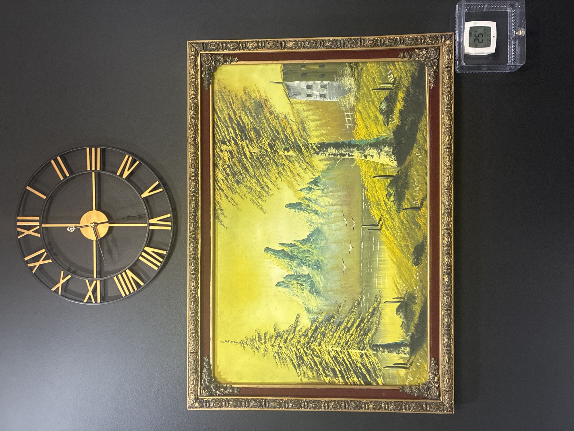 The media file displays an interior scene against a dark grey, textured wall in Jesús del Monte, Mexico. On the left, a large circular wall clock features a black frame with prominent gold Roman numerals and gold hour and minute hands, indicating the time is approximately 8:40.
To the right of the clock, a vertically oriented painting is displayed within an ornate, gold-colored frame adorned with intricate carvings. The painting itself is an abstract or impressionistic landscape, predominantly rendered in shades of vibrant yellow and green. It depicts a flowing river or body of water flanked by dense, stylized trees or foliage, characterized by distinct upward brushstrokes. In the upper left portion of the artwork, a grey, building-like structure with visible windows is present, and faint bird-like shapes can be seen in the upper sky area.
In the upper right corner of the wall, a small, white digital thermometer and hygrometer is encased in a clear, ribbed plastic protective shell. Its digital display shows a temperature of 25.2°C and a humidity level of 52%. The overall scene combines decorative elements with practical measurements, set against a minimalist backdrop.