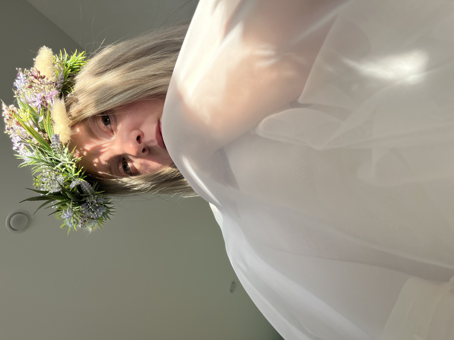A person with fair skin and blonde hair looks down at the camera, wearing a floral crown of greenery and purple flowers. Light streams across their face, creating dappled shadows. A large, semi-transparent white fabric drapes over the right side of the frame, partially obscuring the view. The background is a muted, light gray. The composition is angled, adding a dynamic perspective.