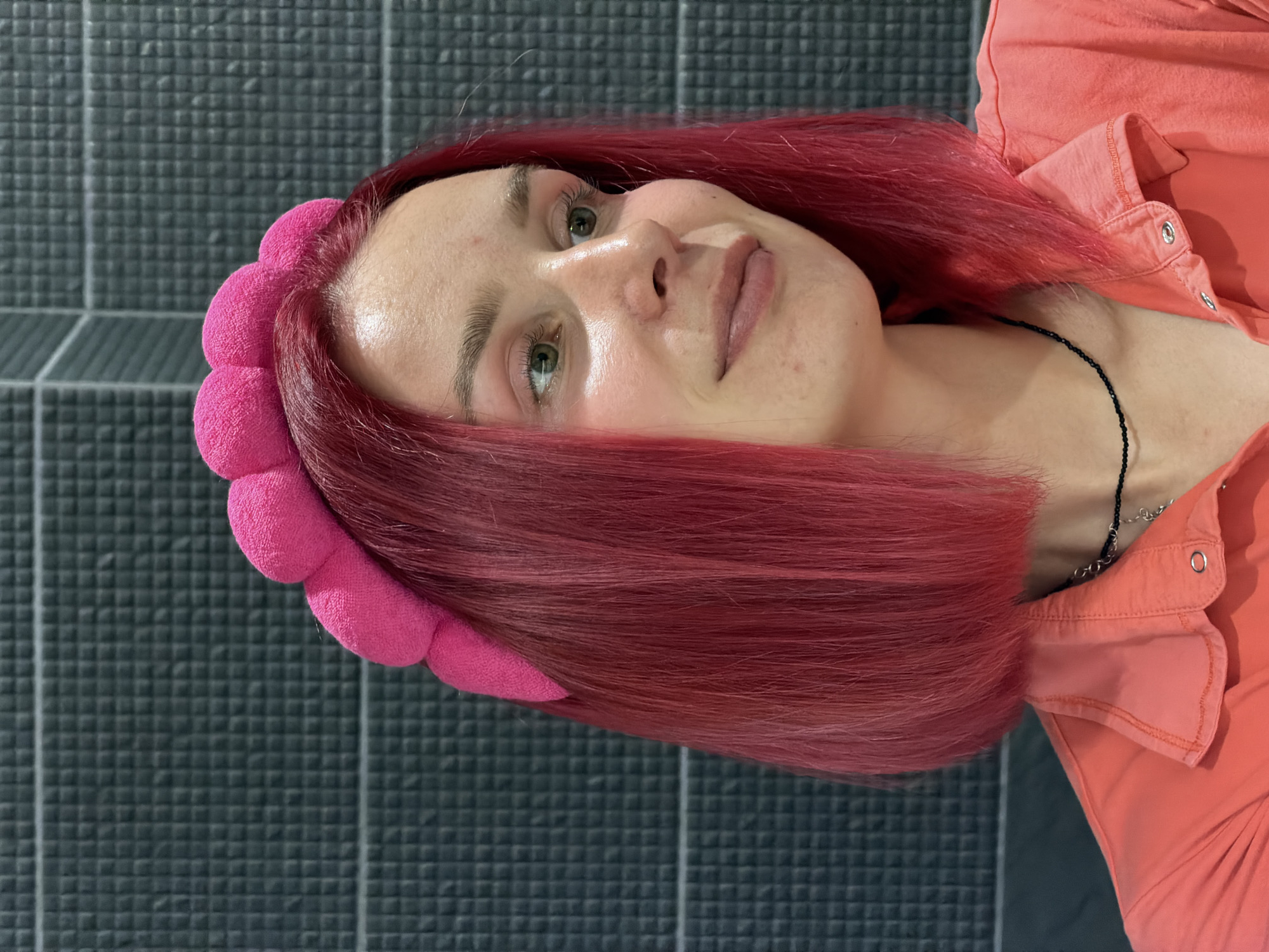 A young adult female is depicted in a head-and-shoulders portrait. She has chin-length, bright pink hair, styled straight, and is wearing a prominent, thick, bright pink headband with a segmented, plush design across the top of her head. Her skin appears fair with some natural texture and slight sheen on the forehead and nose. Her eyes are light green, and she has naturally light eyebrows. She exhibits a subtle, gentle smile while looking towards the right side of the frame. She is dressed in a coral or light orange collared shirt, featuring visible silver snap buttons near the neckline. Around her neck, a thin black beaded choker necklace is visible, layered with a delicate silver chain necklace. The background consists of a dark grey or dark teal wall covered in small, square textured tiles arranged in a grid pattern. A distinct horizontal grout line or divider is present across the mid-section of the tiled wall. The scene is set indoors.