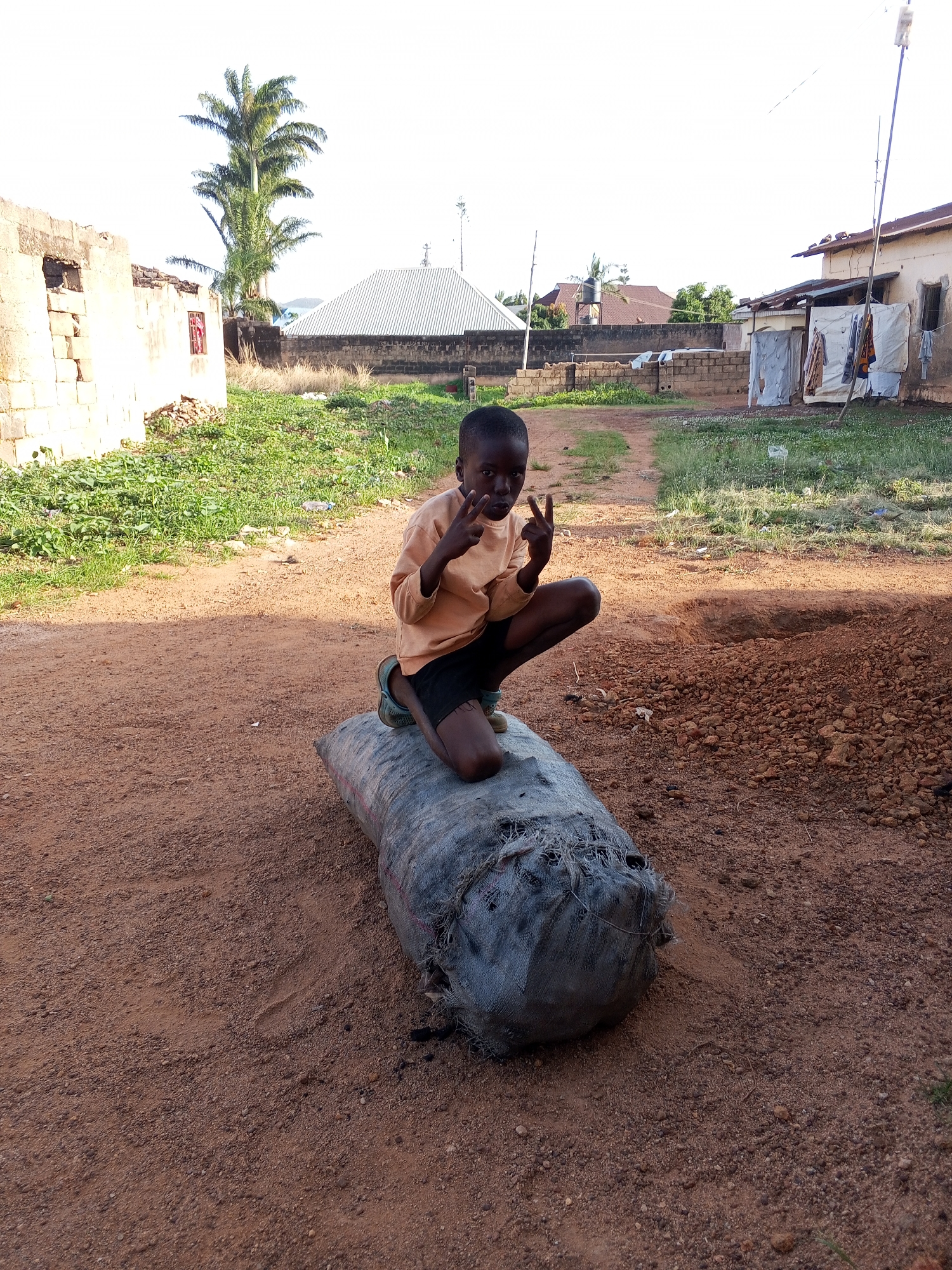 The scene depicts a young boy crouching atop a large, gray, possibly woven bag lying on a reddish-brown, dirt-covered ground, likely a street or yard. He is wearing a light-orange, long-sleeved shirt and dark shorts and is giving the "peace" sign with both hands. The location appears to be Bukuru, Nigeria, based on the style of buildings, vegetation, and the color of the soil.
In the background, there are several one-story buildings, some with corrugated metal roofs, and patches of grassy vegetation. A tall palm tree is visible in the mid-ground. Clothes appear to be hanging outside one of the buildings. The light suggests it is daytime. The ground surface around the boy is primarily dirt with pebbles, and appears uneven.