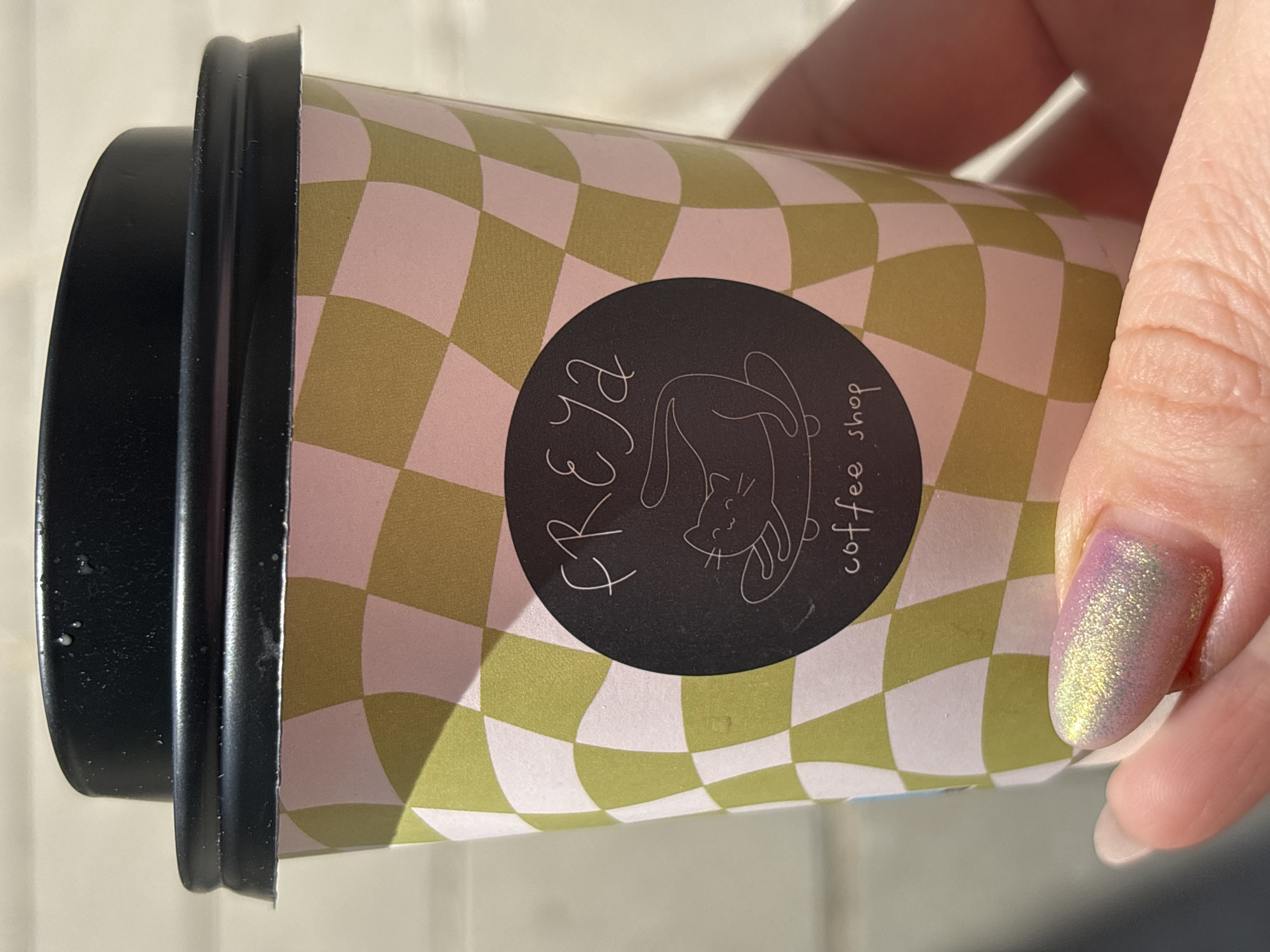 A close-up, angled shot depicts a paper coffee cup held in someone's hand. The cup has a black plastic lid and a checkered pattern of light green and light pink squares. A black circular logo on the cup features the word "Freya" in script lettering above a cartoon illustration of a cat, and the words "coffee shop" in small print to the side of the cat. The person's thumb is visible, adorned with iridescent, color-shifting nail polish. While the coffee cup is from a coffee shop called Freya, the image does not provide information on the location or the city of Sofia, Bulgaria.