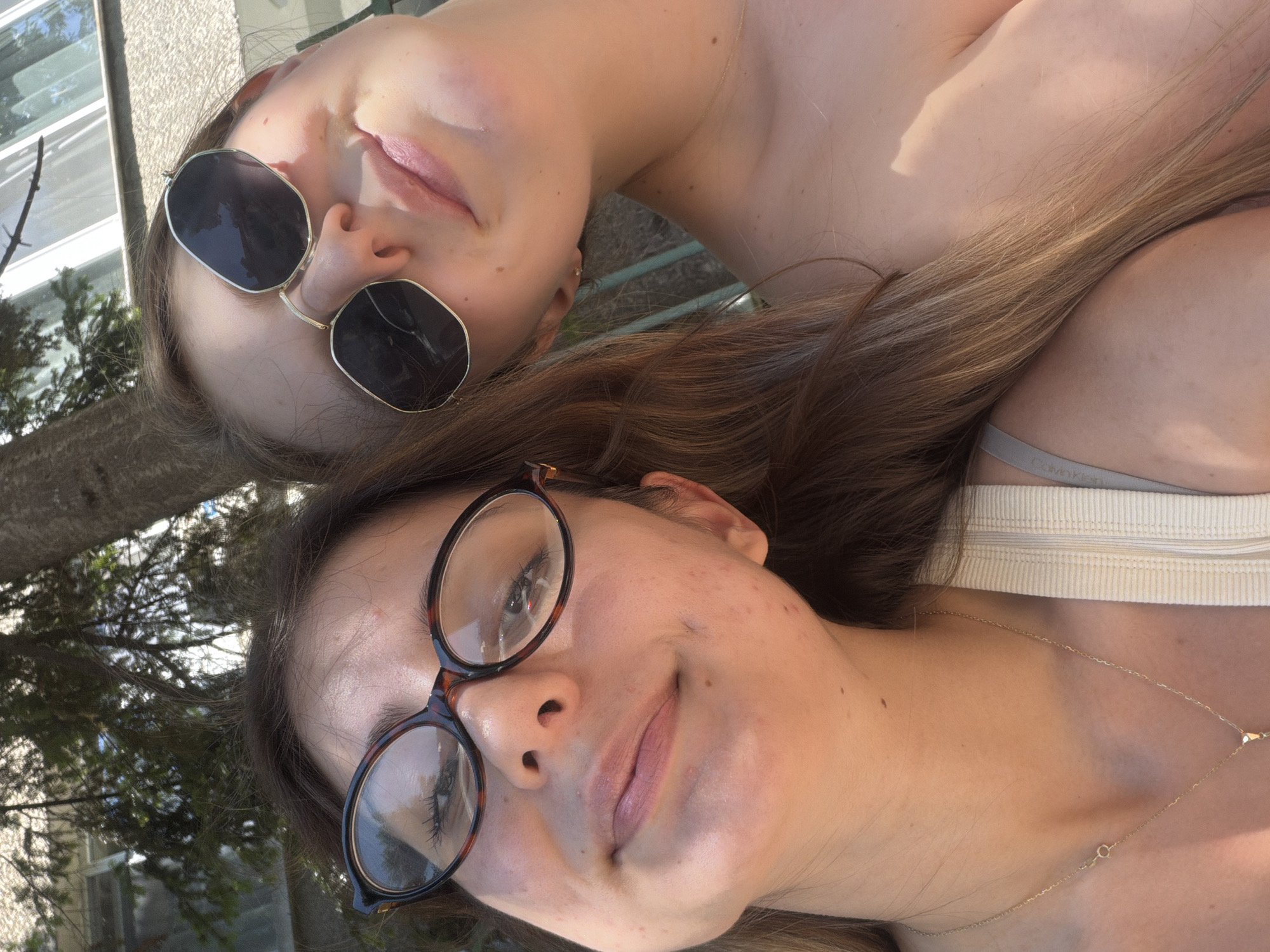 In an outdoor, sunlit setting, two young women are captured in a close-up, selfie-style photograph. The woman on the left, wearing tortoiseshell glasses, faces the camera with a subtle smile. Her skin shows some blemishes, and her dark brown hair frames her face. The woman on the right, partially obscured, wears hexagonal sunglasses and her face is tilted upwards. Her fair hair is visible. They appear to be in a relaxed, social environment, possibly a park or a garden, with a hint of a building and foliage in the background. The image evokes a sense of leisure and friendship. There is no specific indication of the city of Burgas, Bulgaria, within the visual information provided.