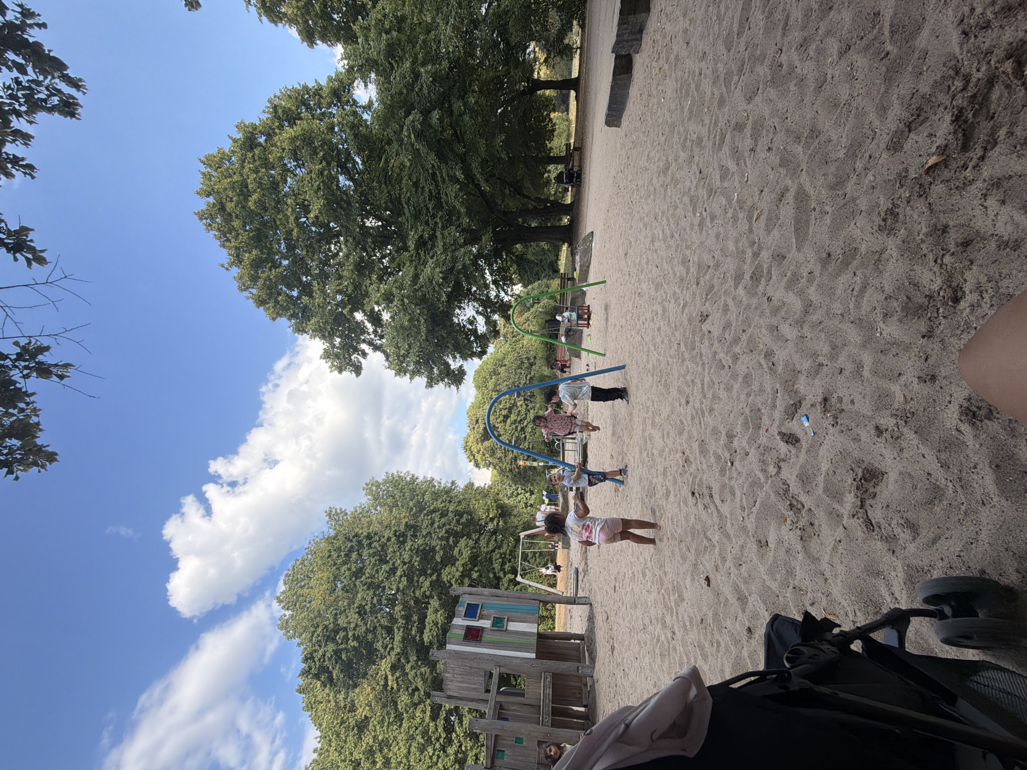 This is a photograph taken at the playground in the Landschaftspark Duisburg-Nord, Germany. The image captures a sunny day with a blue sky and scattered white clouds. In the foreground, a black stroller is partially visible on a sandy playground surface. Several children are present. One girl in a pink top and shorts is standing on the sand. Another child is on a swing. A boy in a striped shirt and blue shorts is near the swing set, and another child in a patterned shirt is also on the swing. In the background, a wooden playground structure is visible, along with more trees and park amenities. The overall impression is of a lively, outdoor recreational space.