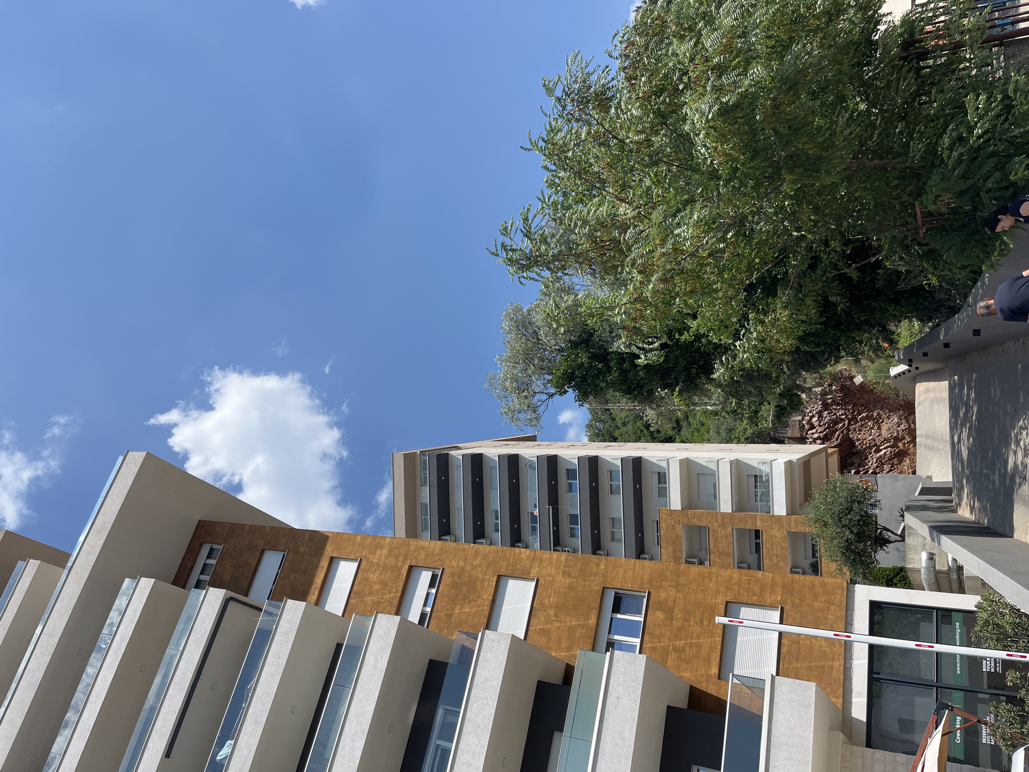The image shows a contemporary apartment building in Budva, Montenegro, under a clear blue sky with scattered clouds. The building features modern architecture with white balconies and warm brown facade panels. To the right of this building, another taller apartment complex is visible, also with balconies. In the foreground, a pathway leads uphill, flanked by a concrete railing. Lush green trees and vegetation are present on the right side of the pathway, with a rocky, wooded hillside in the background. Two individuals, an adult male and an adult female, are walking up the pathway away from the camera. The scene appears to be in a residential or tourist area, likely during daytime.
