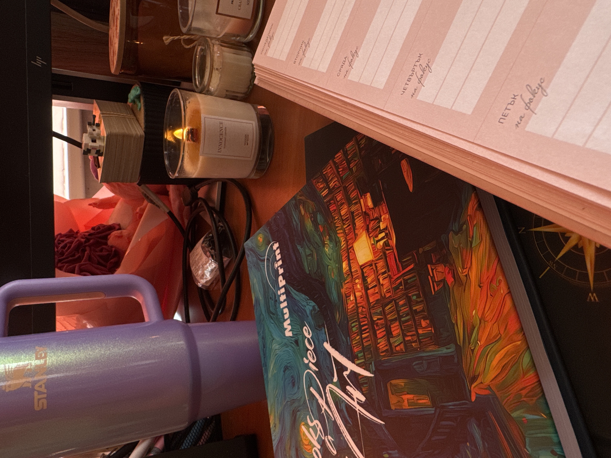 This image depicts a desk scene in Kostinbrod, Bulgaria. On the left is a tall, iridescent purple Stanley tumbler with a handle. To the right of the tumbler are various objects arranged on a wooden surface. A lit candle with the label "INNOCENCE scented candle" is prominent, alongside two smaller glass jars, one appearing to contain a candle or wax. Behind the candle is a stack of what appears to be small, light-brown blocks, possibly toys or decorative items, bound by a rubber band. A portion of a computer monitor is visible in the background. In the foreground, a book with a title that includes "MultiPrint" and stylized text is laid open, showcasing a vibrant illustration of a library with warm lighting. Next to the book is a pink planner with handwritten entries in Bulgarian, including days of the week like "ПОНЕДЕЛНИК" (Monday), "ВТОРНИК" (Tuesday), "СРЯДА" (Wednesday), "ЧЕТВЪРТЪК" (Thursday), and "ПЕТЪК" (Friday), each followed by the phrase "на фокус" (on focus). A compass rose design is partially visible on a dark surface beneath the planner and book. The overall environment suggests a workspace or study area.