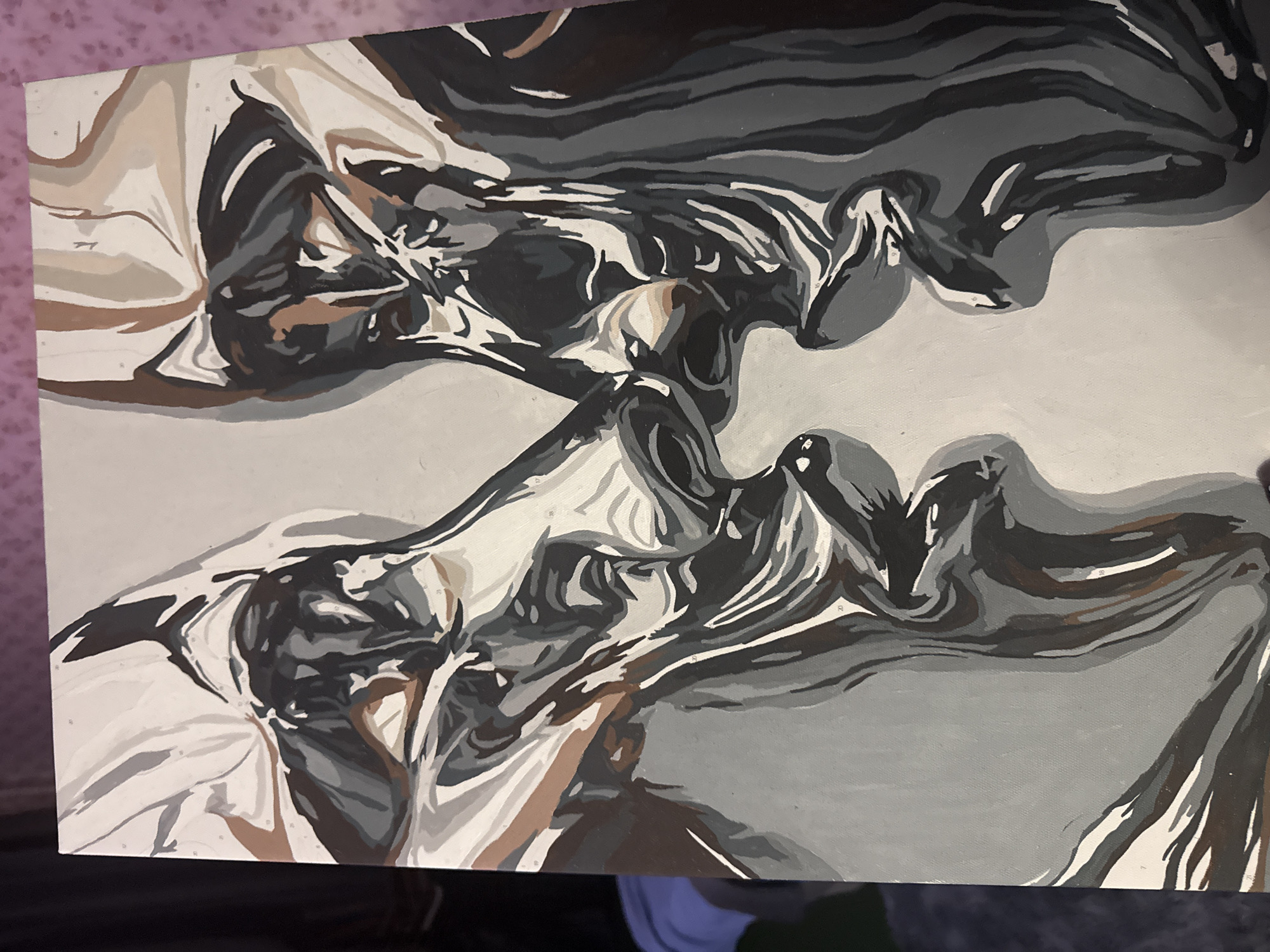 A close-up photograph shows an unfinished paint-by-numbers artwork. The painting consists of grayscale tones and earth-toned hues. Swirling, abstract patterns are depicted in the upper and lower regions of the canvas. The location is not discernible from the image. There are no people in the frame. The environment appears to be indoors.