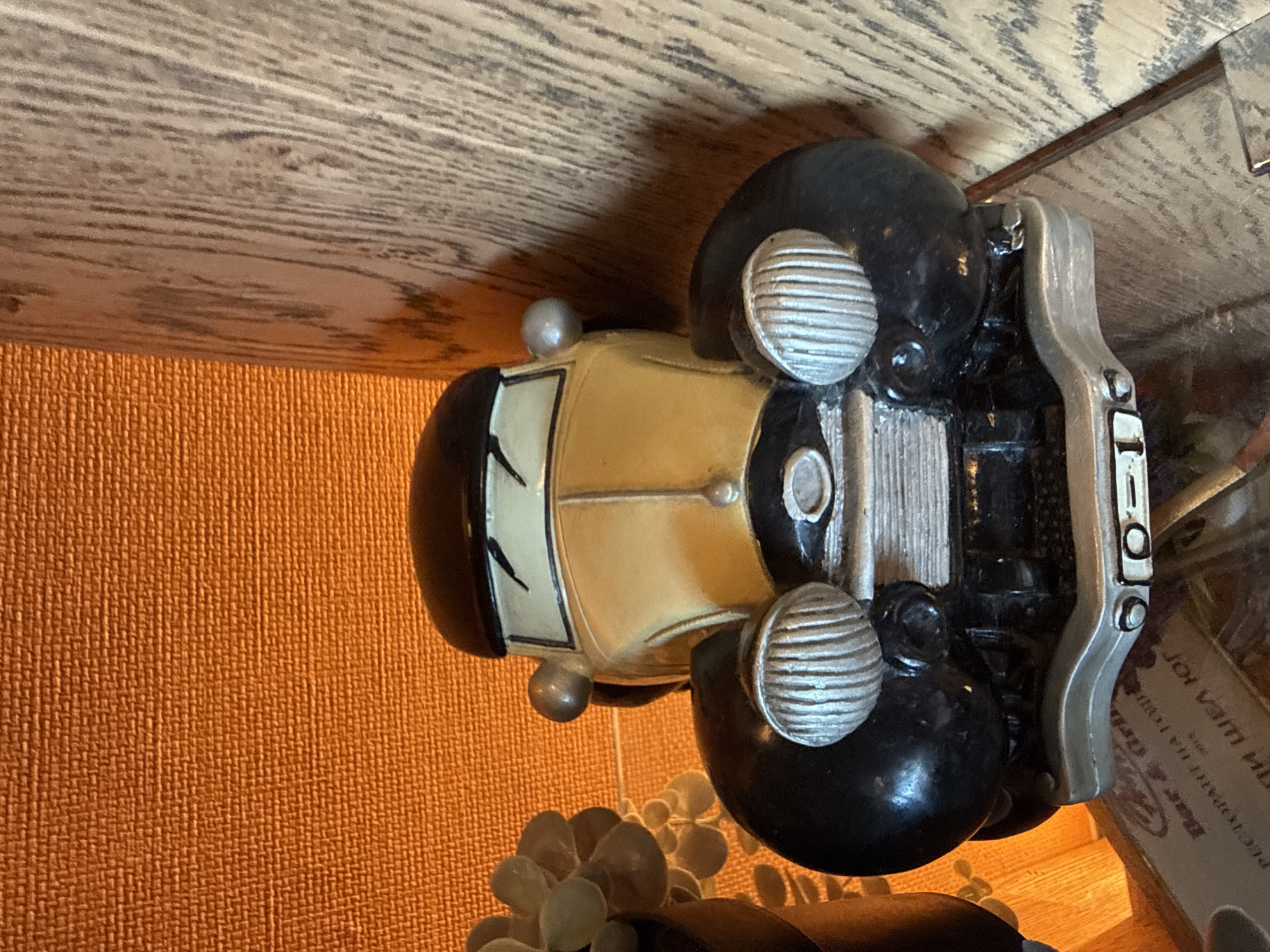 The image displays a close-up, top-down view of a decorative model car, oriented vertically with its front pointing upwards. The car, styled like a vintage automobile, has a light yellow or beige body, black fenders, and a black roof. Its most prominent features include large, circular silver headlights with horizontal grilles, a silver front grille with vertical slats, and smaller silver dome-shaped lights on the front fenders and possibly the roof. The windshield has black stylized markings, potentially representing wipers or a face. The rear bumper and surrounding structure are silver.
The model car rests on an unseen surface, with a textured, burnt orange material forming the background to its left and below. Above the car, a wooden surface with a distinct grain pattern is visible. Below the front of the car, out of focus, are parts of what appear to be artificial succulent plants or similar decorative foliage. To the right of the car, a blurry brown object or box shows partially visible text, largely in Cyrillic script. Phrases such as "Bar & Grill" and "РЕСТОРАНТ НА ТОЧНО" (Restaurant exactly/precisely) can be deciphered, along with other incomplete words like "ЖОИ ЯМ" (Joi Yam). The scene is static, depicting a collection of decorative items in what appears to be an indoor setting.