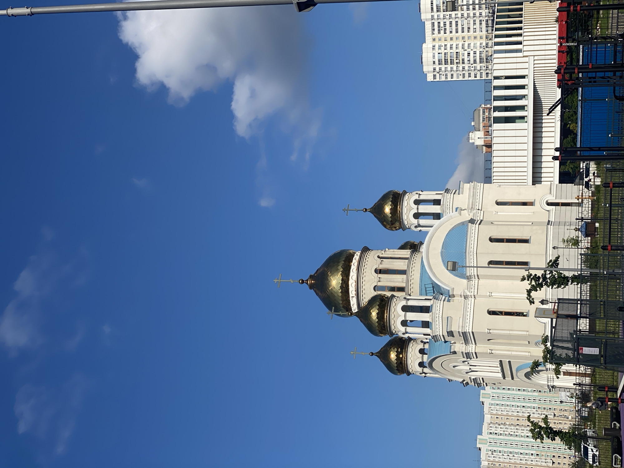 A low-angle outdoor shot captures a white Orthodox church with golden domes under a clear blue sky with scattered clouds. The church has multiple domes topped with crosses, and blue accents on its roofline. In the foreground, a dark metal fence surrounds a playground area with visible structures, including a blue container and dark poles. Behind the church, residential buildings of varying heights are visible. The lighting suggests it is daytime.