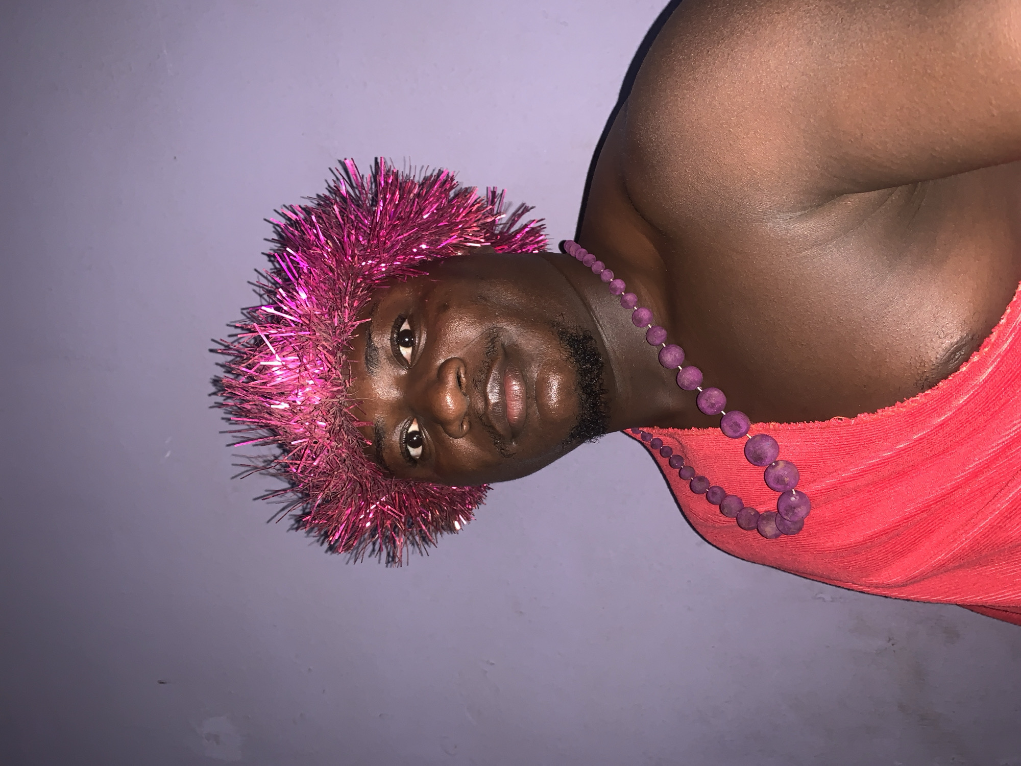 A medium shot shows a dark-skinned man wearing a bright pink, spiky tinsel crown and a purple beaded necklace. He has a goatee and is looking at the camera with his mouth slightly open. He is draped in a bright red fabric that covers his shoulder and chest. The background is a solid, muted purple wall with some subtle texture. The image appears to have been taken indoors in Akwanga, Nigeria, likely in a residential setting, as indicated by the plain wall and the intimate framing of the subject.