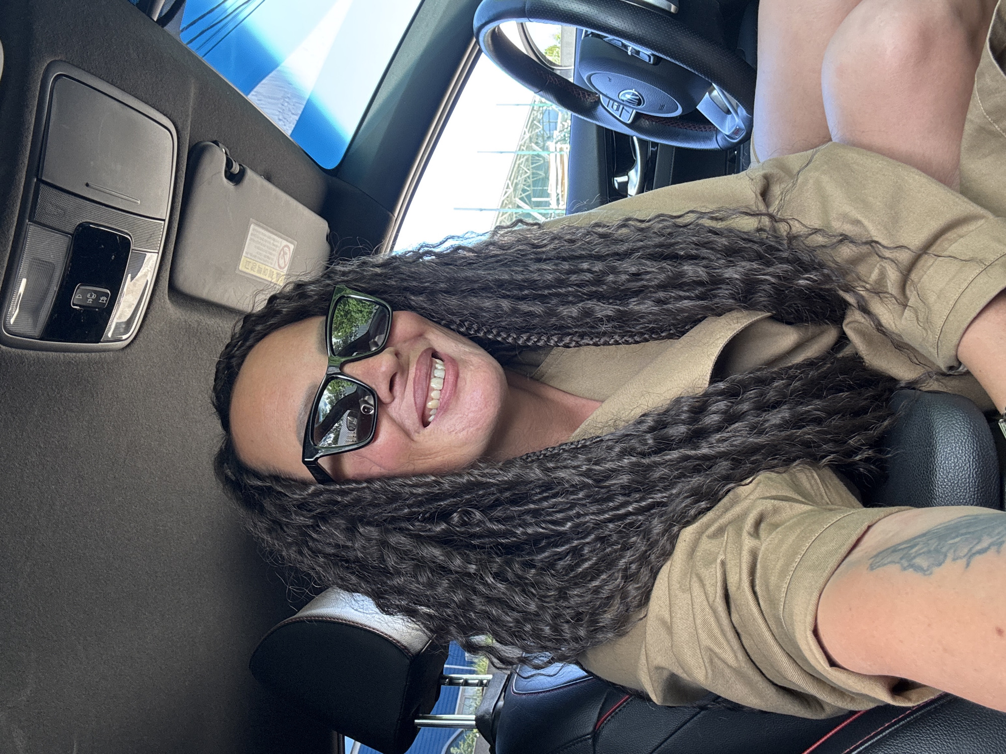 A medium-shot, upward-angled view depicts a woman inside a car. The woman, wearing sunglasses and a tan shirt, smiles towards the camera. Her long, dark, curly hair is loosely styled. She appears to be seated in the driver's seat. The interior of the car is visible, including the steering wheel and part of the dashboard. Through the car windows, a partial view of an outdoor scene is visible. The location is identifiable as Chaikovsky, Russia.