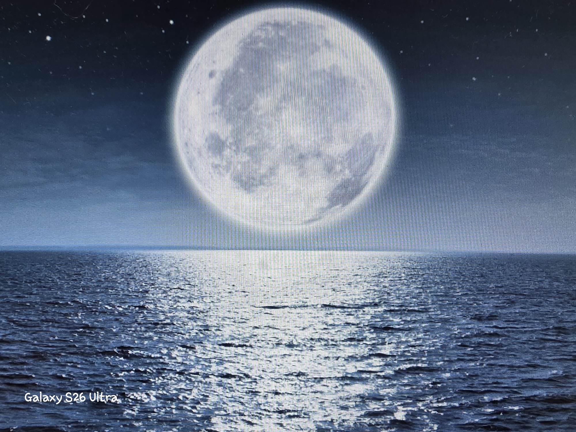 The image depicts a serene night scene with a large, luminous full moon dominating the upper half. The moon is detailed, showing craters and texture, and is set against a dark blue sky dotted with small, distant stars. Below the moon, a calm body of water stretches to the horizon, reflecting the moonlight in a shimmering path. The water is dark blue, with small waves catching the light. In the bottom left corner of the image, the text "Galaxy S26 Ultra" is visible. The scene is a digital rendering or photograph of a natural landscape, not depicting any specific location within the city of Plovdiv, Bulgaria. There are no people, actions, or interactions present in the image.