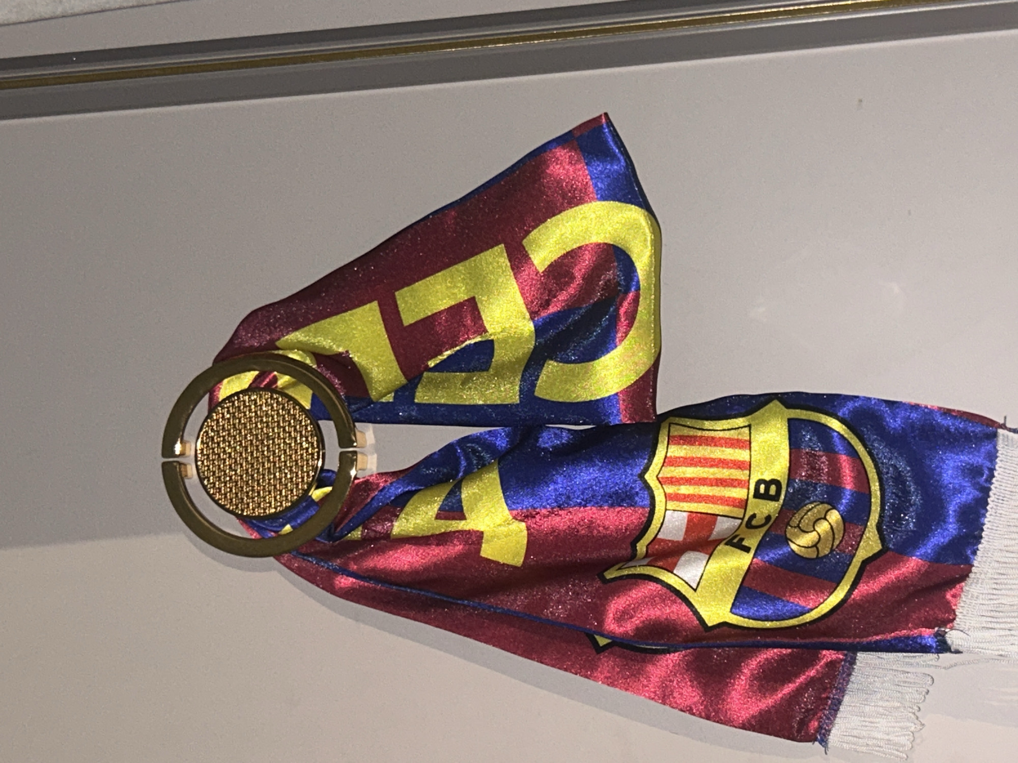 The image shows a FC Barcelona scarf displayed indoors. The scarf is predominantly red and blue with yellow detailing, featuring the FC Barcelona crest and the word "FCB". It is draped through a gold-colored circular object with a textured center. The background appears to be a plain, light-colored wall. The location, Topolovgrad, Bulgaria, is not visually evident in the image.