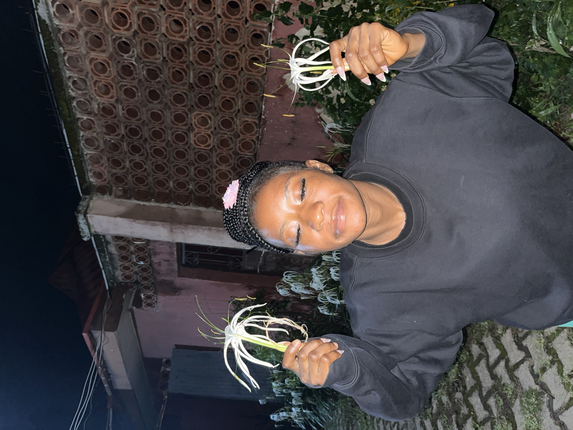 A young Black woman is standing outside at night, holding two white spider lilies. She is wearing a black sweatshirt and has her eyes closed and her mouth slightly open, appearing relaxed. A pink flower is tucked into her braided hair. Behind her, a pink building with decorative brickwork is visible, along with lush green foliage. The ground in front of her is paved with interlocking bricks and has patches of grass. The location is identified as Jos, Nigeria.