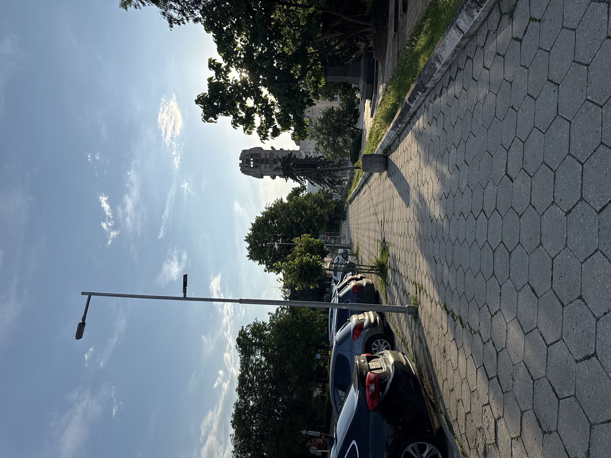 This image, taken in Shumen, Bulgaria, depicts an outdoor urban or park scene, presented in a sideways orientation. A long sidewalk paved with grey hexagonal interlocking pavers stretches from the foreground towards the background on the right. Patches of grass grow along the edges of the sidewalk.
Several dark-colored cars are parked in a row along the left side of the street next to the sidewalk. A tall, slender grey street lamp post with two horizontal light fixtures stands prominently in the midground. Dense green trees line both sides of the street, casting long shadows across the sidewalk and parked cars.
In the background, partially visible through the trees, is the distinctive, angular structure of the Monument to 1300 Years of Bulgaria. The sky is bright blue with scattered white clouds. The sun, high in the sky, is visible through the tree canopy on the upper right, casting strong sunlight and indicating a late morning or early afternoon. The weather appears clear and sunny. No people or visible text are present in the image.