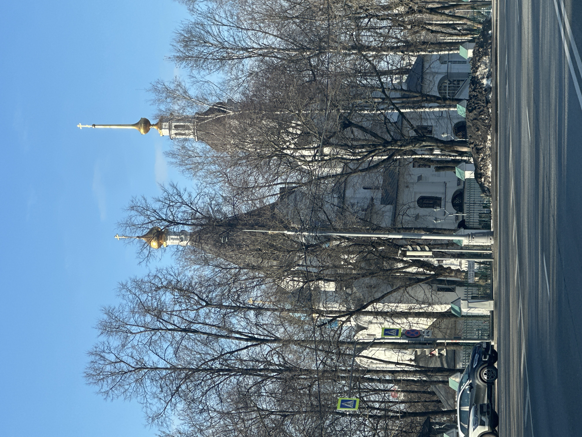 The image shows a street view in Moscow, Russia, featuring the Danilov Monastery. The architecture of the monastery is visible, with its distinctive towers topped with golden domes and crosses, partially obscured by bare trees lining the street. The sky is clear and blue. A paved road runs along the right side of the image, and parked vehicles are visible. Pedestrian crossing signs are also present. The scene appears to be captured during winter or early spring, as the trees are devoid of leaves.