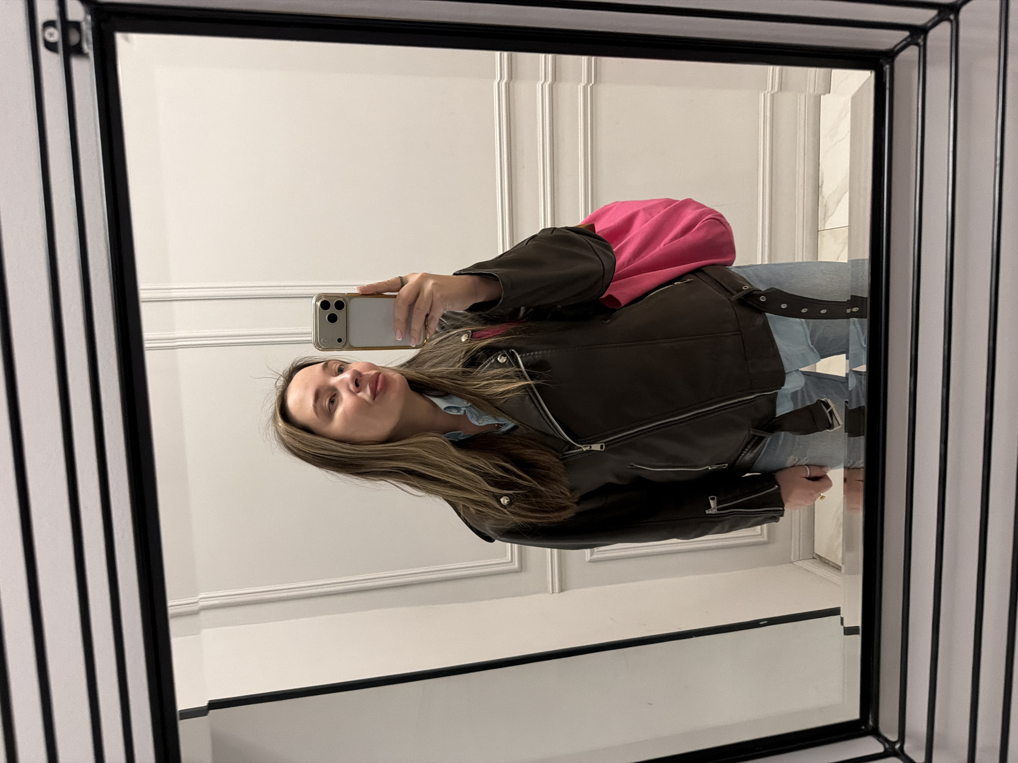 A person stands in front of a mirror taking a selfie, holding a smartphone. The individual is wearing a brown leather jacket and jeans with a pink bag over their shoulder. The mirror has a black frame and is set against a white wall with decorative paneling. Based on the style of architecture, this could be a bathroom or dressing room in a location in Moscow, Russia.