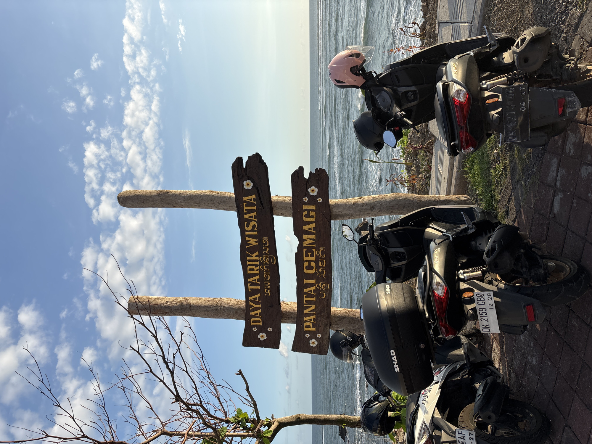 The image shows a scene near Pantai Cemagi in Mengwi, Indonesia. A wooden sign with the Indonesian words "DAYA TARIK WISATA PANTAI CEMAGI" is prominently displayed. Several motorcycles are parked near the sign, with one having a black SHAD storage box and another with a pink helmet placed on it. The license plate of a motorcycle is visible, reading "DK 2159 GBO 12 28." In the background, the ocean can be seen under a cloudy sky. A tree branch extends into the frame on the left side.