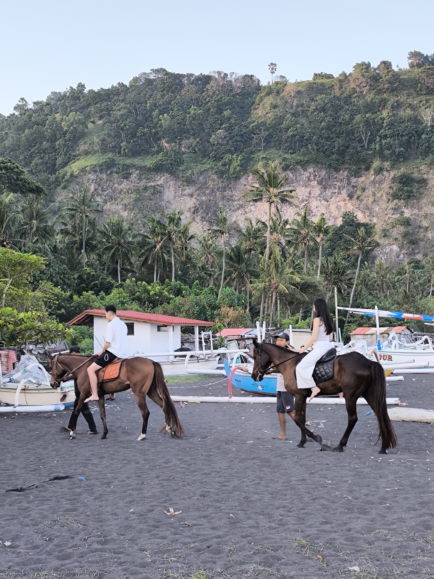 Two people are riding horses on a black sand beach in Karangasem, Indonesia. The rider on the left is a person wearing a white shirt and black shorts. The rider on the right is a person wearing a white tank top and white pants. Both horses are brown. In the background, there are palm trees and a cliff. There are also several boats on the beach. The scene appears to be a tourist attraction.