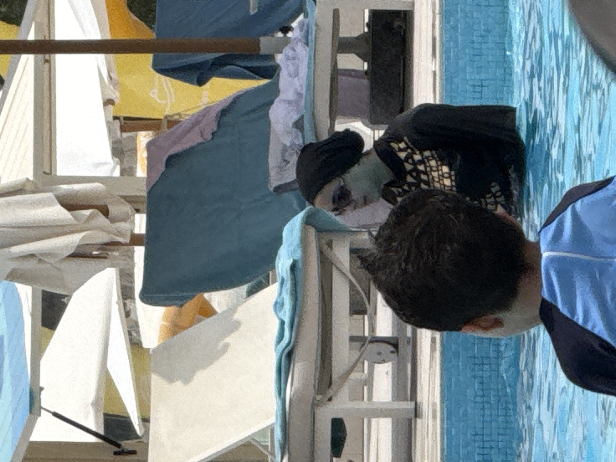 The image depicts a poolside scene at a resort in an unspecified location in the United Arab Emirates. In the foreground, the back of a child wearing a blue and black swimsuit is visible, facing the pool. In the middle ground, a woman in a black burkini and sunglasses is partially submerged in the pool, with a light blue mask on her face. She is wearing a patterned top over her burkini. The background features several lounge chairs with blue towels and white umbrellas, suggesting a resort or hotel setting. The water in the pool is a clear blue, and the overall lighting indicates it is daytime.