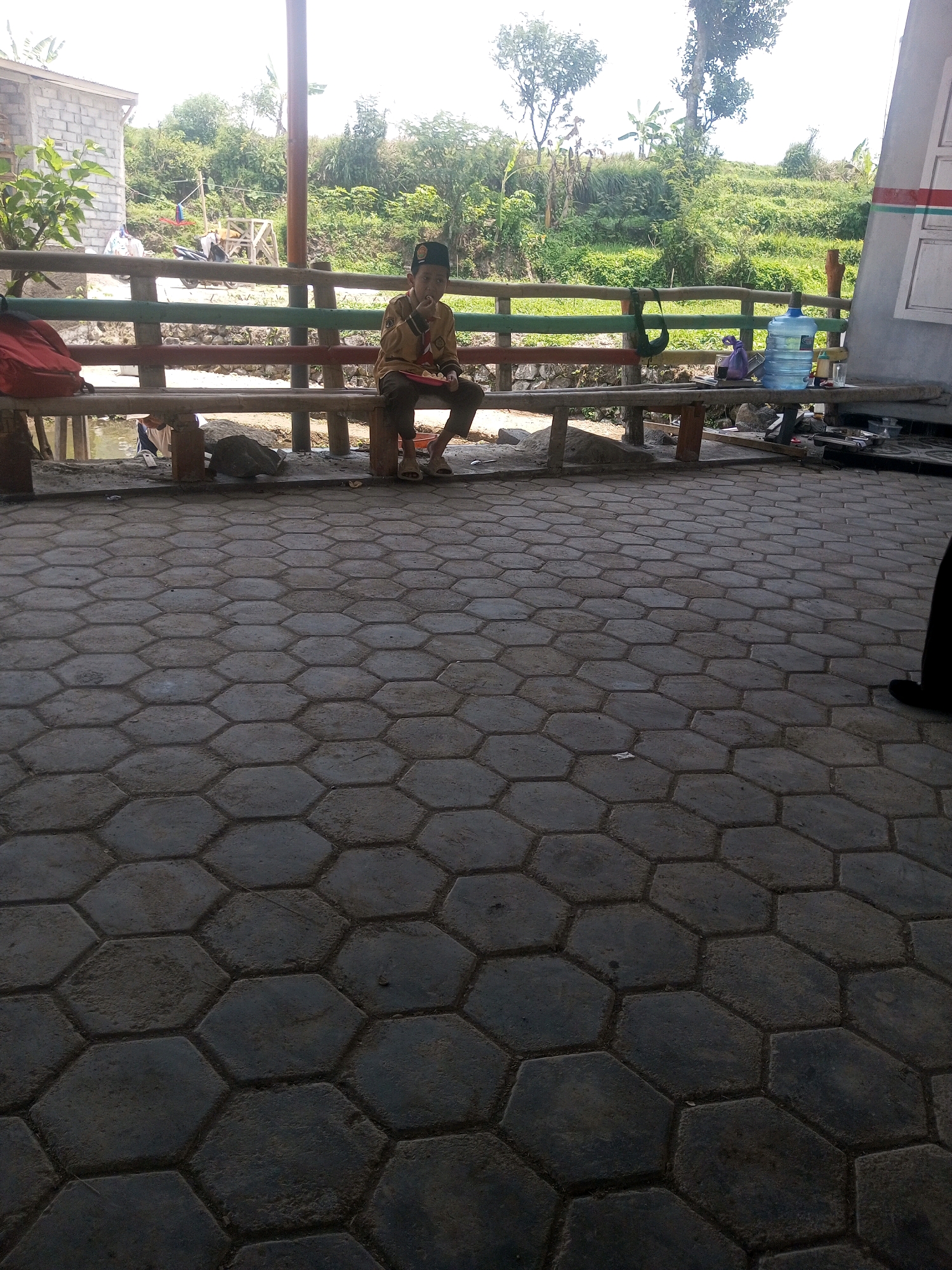 In Kalibeber, Indonesia, a young person wearing a brown uniform and a black cap sits on a wooden bench, eating. The bench is located outdoors on a paved area covered with hexagonal tiles. Behind the bench, a wooden railing separates the paved area from a natural landscape with lush green trees and vegetation. In the background, a motorcycle and a small wooden structure are visible. To the right of the bench, a large blue water dispenser and a glass are placed on a table. The overall scene appears to be a casual outdoor setting, possibly a rest stop or a recreational area in the vicinity of Kalibeber.