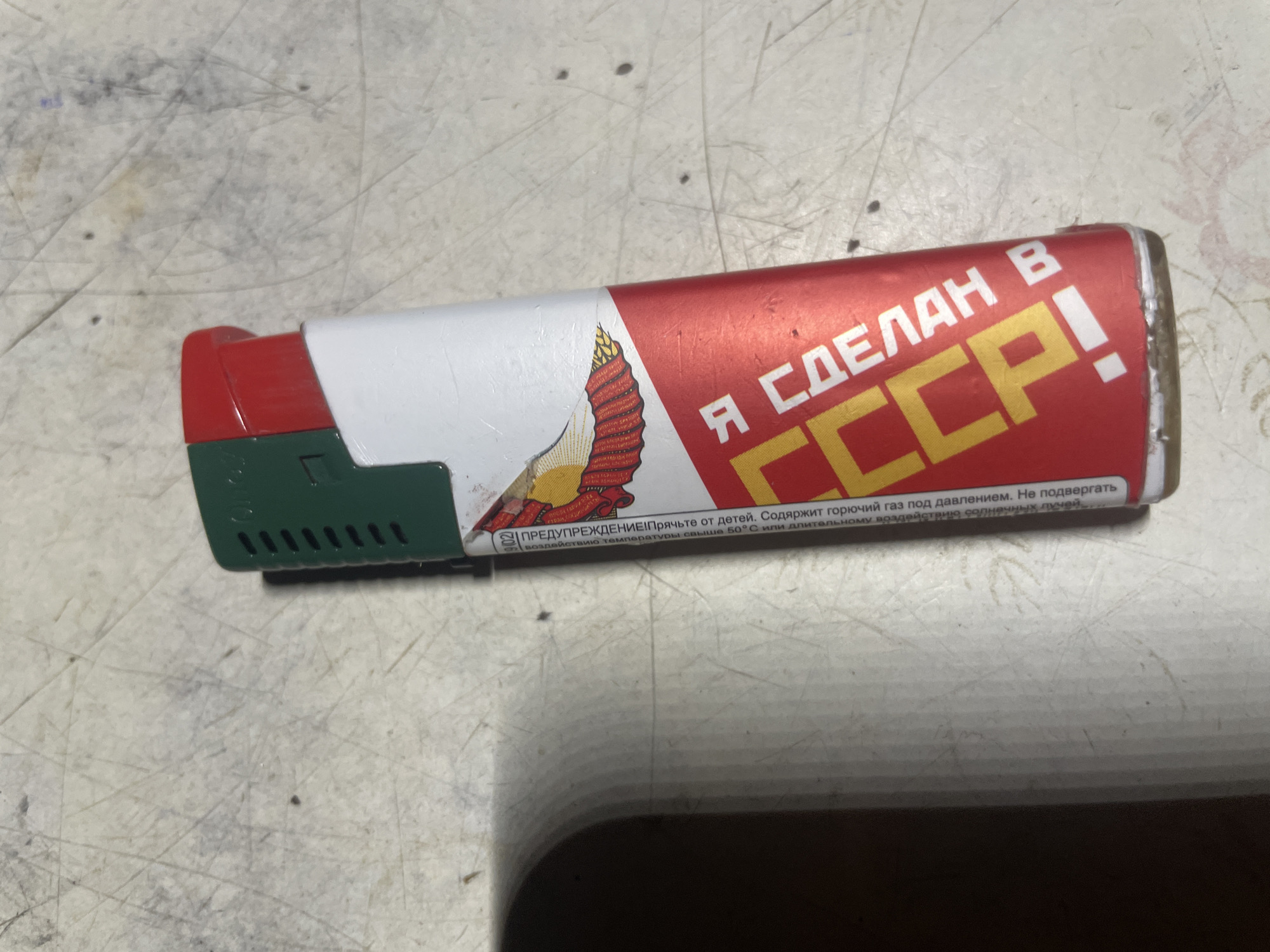 The image shows a close-up, top-down view of a red, white, and green disposable lighter with "Я СДЕЛАН В СССР!" (I AM MADE IN THE USSR!) printed in yellow and white text on a red background, along with a graphic of a bird with red and yellow wings. The lighter is positioned diagonally on a light-colored, textured surface that appears to be a workbench or table with scuff marks and some scattered dark specks. The surface is lit from above, casting a shadow to the left of the lighter. The background is out of focus. There is no indication of the city of Laishevo, Russia, or any human presence in the image.