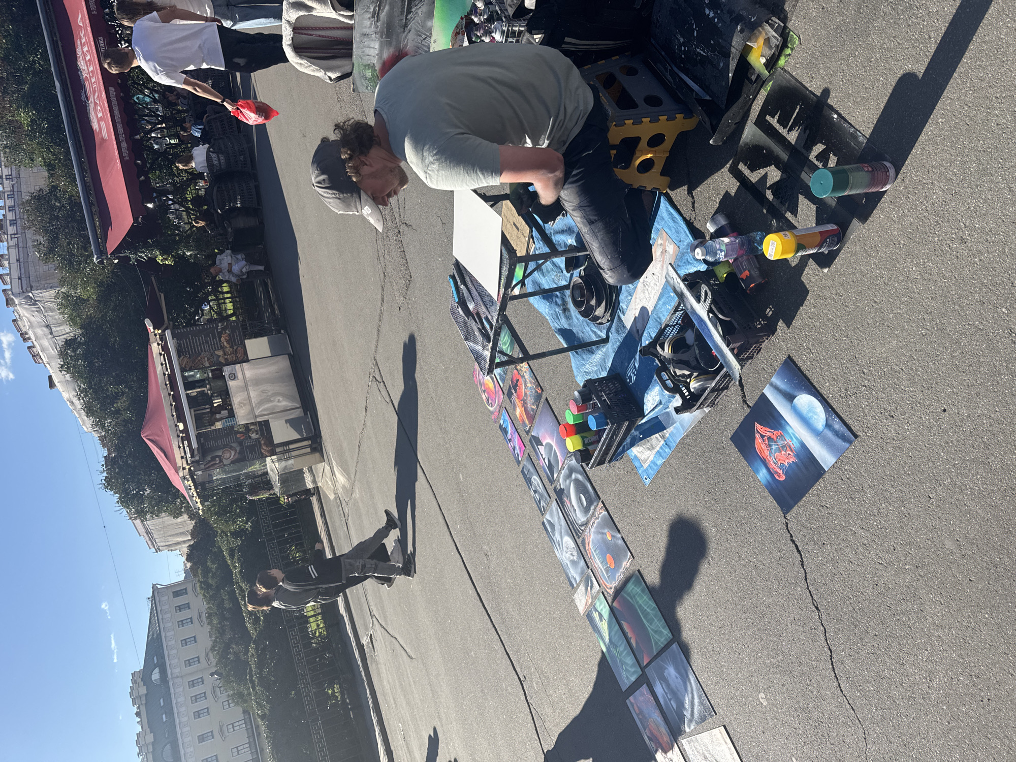 An outdoor scene in Saint Petersburg, Russia, features a street artist creating art on the asphalt. The artist, a man wearing a baseball cap and a light-colored t-shirt, sits on a yellow folding stool behind a small table. Several spray paint cans and stencils are laid out on the ground around him. A collection of finished artwork, likely spray-painted pieces, is displayed in a line on the asphalt in front of him, creating a vibrant showcase. In the background, buildings with distinctive architecture and trees line the street, suggesting an urban park or public square setting. Two individuals walk in the background, adding a sense of pedestrian activity to the scene. The weather appears to be sunny with a clear blue sky.