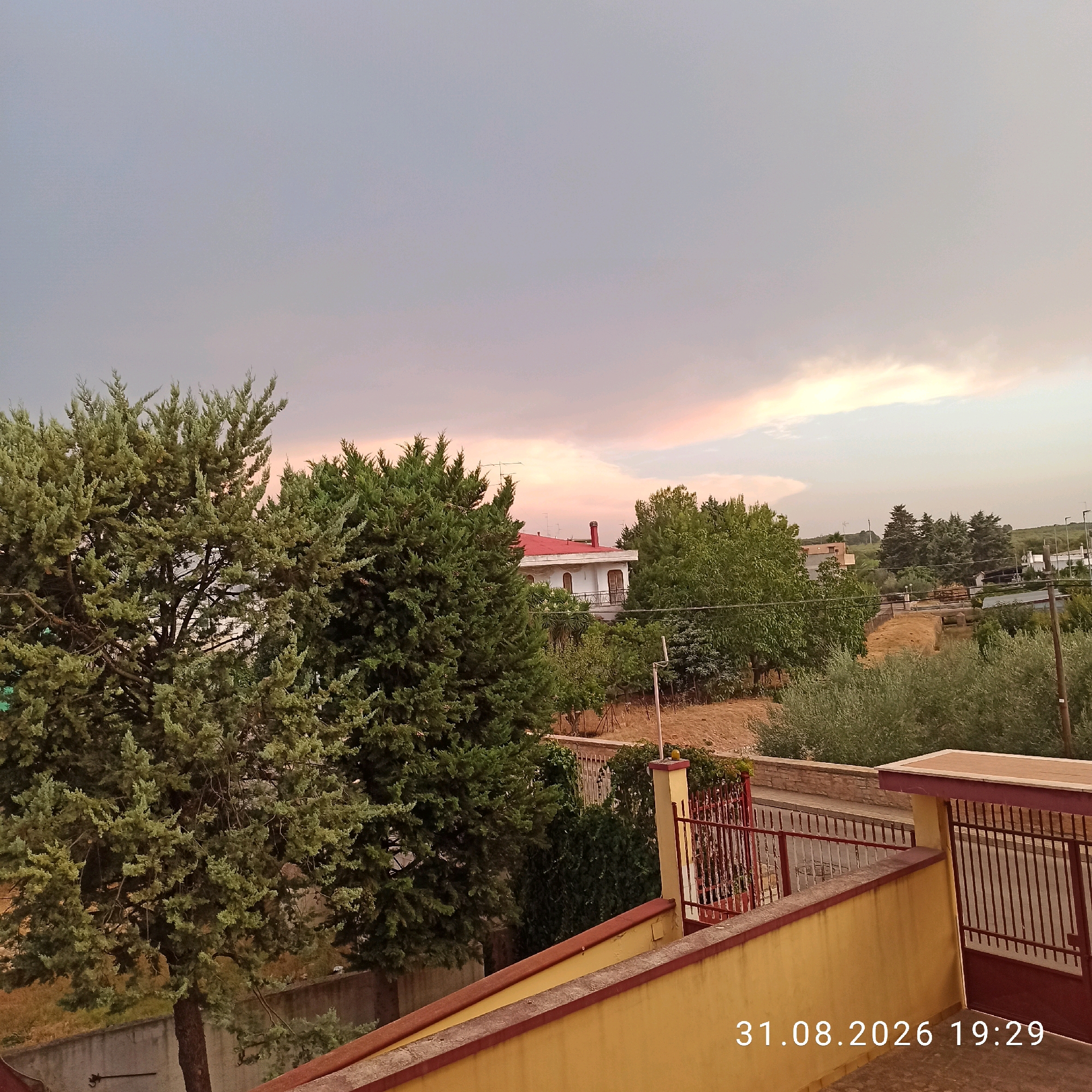 The image shows a view from a balcony overlooking a residential area with vegetation, a building, and a fence. The sky is partly cloudy with hues of pink and orange, suggesting either sunrise or sunset. In the foreground, there are trees with green foliage. A building with a red roof and balconies is visible in the middle ground, surrounded by trees. A red metal fence with a gate is also present. The time stamp on the image reads "31.08.2026 19:29". The scene is likely a residential property in Bitonto, Italy, based on the timestamp and typical Mediterranean architecture and vegetation. There are no people visible in the image.