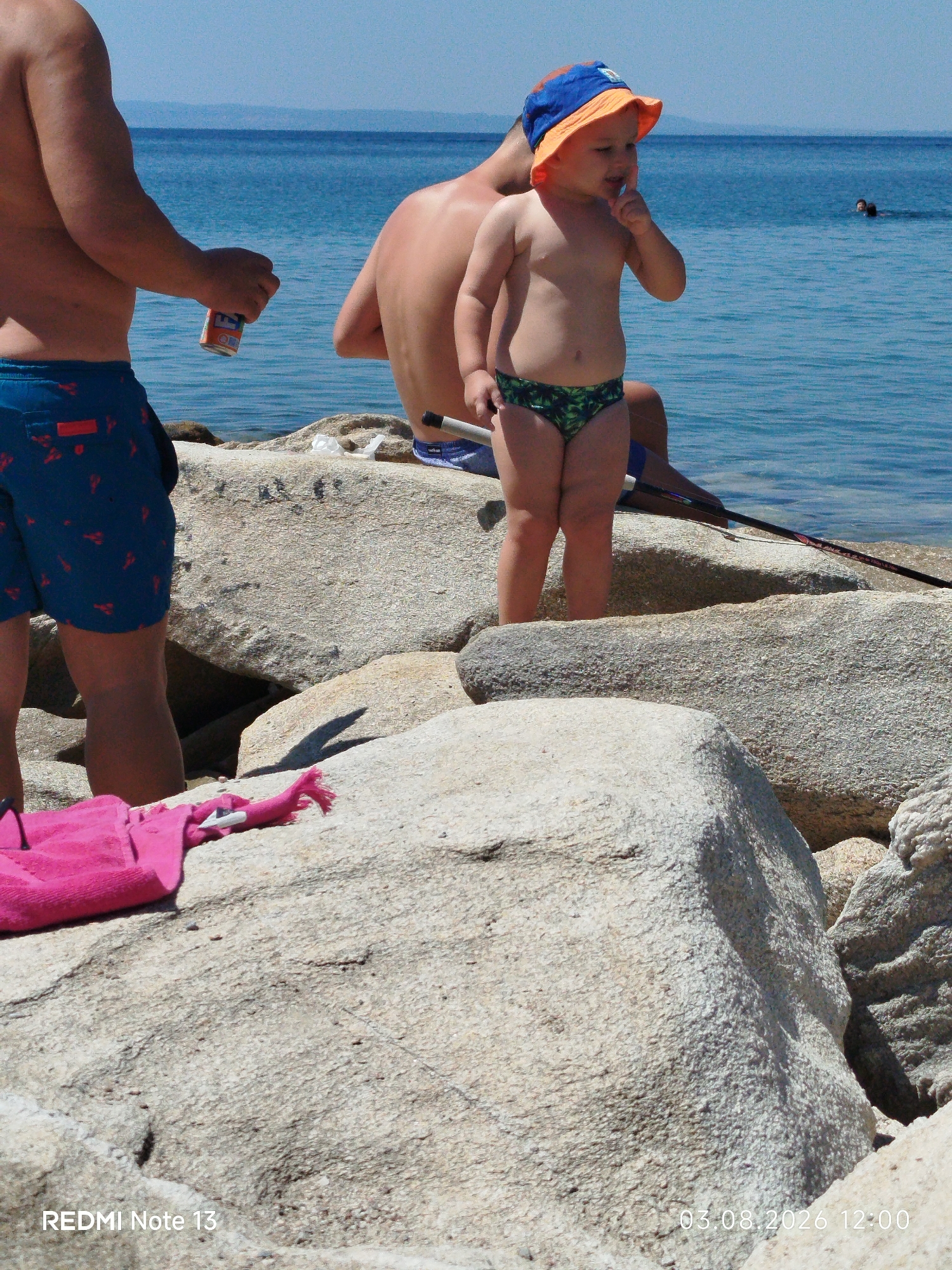 The image depicts a sunny day at a rocky beach, likely in Sithonia, Greece, given the timestamp. In the foreground, a child wearing a blue and orange hat and green and black patterned swim trunks stands on large rocks. The child has their hand to their mouth, appearing to be eating something. To the left, an adult's torso and legs are visible; they are wearing blue swim trunks with a red pattern and holding an orange can. Another adult is seated behind the child, with their back to the camera. In the background, the blue sea stretches out, with two people swimming in the distance and a fishing rod extending towards the water from behind the seated adult. A pink towel is draped on the rocks in the lower left portion of the image. The timestamp "03.08.2026 12:00" is visible in the bottom right corner, and "REDMI Note 13" is in the bottom left.