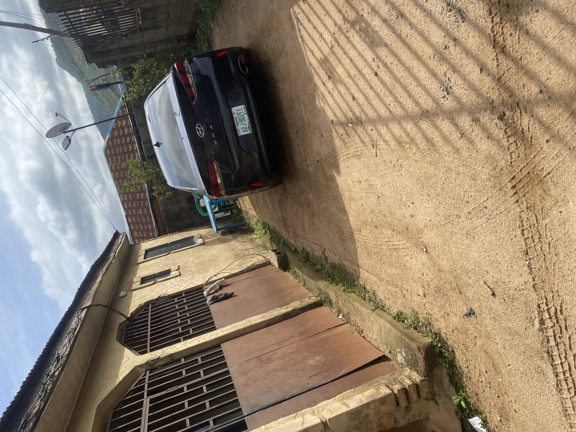 The image displays an unpaved, light brown dirt thoroughfare in a residential area within Abuja, Nigeria. A dark-colored Hyundai Kona sedan, bearing Nigerian license plate FCT ABUJA ABJ-902FE, is parked facing away from the viewer on the right side of the frame. To the left, a single-story, light brown stucco-finished building with a corrugated roof features two prominent arched double metal doors with security grates, and additional windows, some with blue frames. Overhead, electrical wires are strung along the building and across the sky. Small patches of green vegetation grow along the base of the building and near a partially visible blue plastic table and white chair. Further back, a utility pole supports a satellite dish and a street lamp. In the background, a grey concrete block wall topped with a metal fence is visible, alongside another building with a rusted corrugated roof. A distant mountainous landscape is discernible under a partly cloudy sky. Distinct shadows from an unseen structure or vegetation are cast across the dirt ground, which also shows tire tracks. No individuals are present.