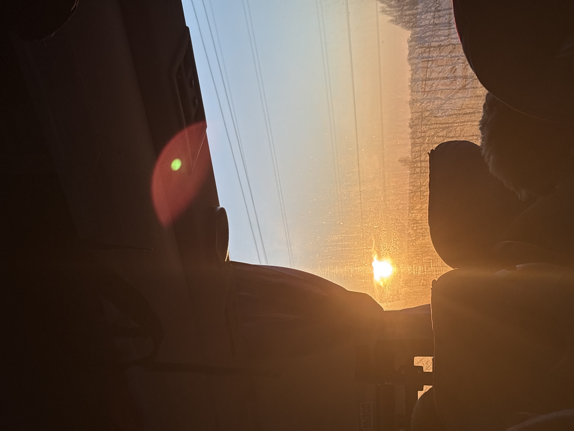 The image shows a view from inside a vehicle, possibly a car, looking out the front windshield towards a bright, low-angle sun. The sun is positioned near the horizon, casting a golden glow through the dusty window. Several power lines are visible against the lighter, upper portion of the sky. Interior elements of the vehicle, like the dashboard, seats, and visor, are silhouetted in the foreground. The location is assumed to be within the city of Osa, Russia, based on user input.