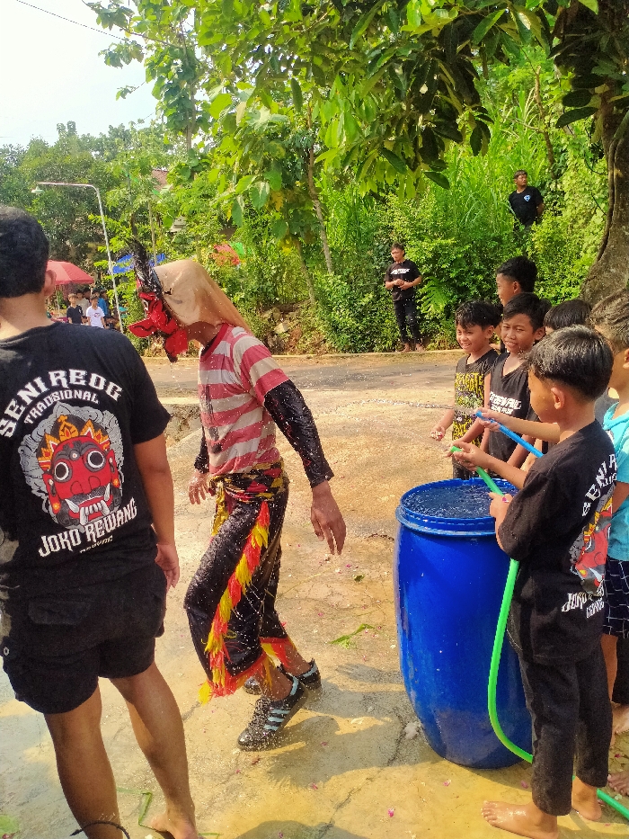 This image depicts an outdoor scene in Gebyog, Indonesia, characterized by a lively interaction involving a masked performer, several children, and other observers.
In the foreground, a person is dressed in a traditional costume featuring a red, demonic-style mask with black accents, a light brown head covering, a red and white striped long-sleeved shirt, and black pants adorned with yellow and red fringes. This individual is wearing black athletic shoes and is notably wet, with visible water droplets on their clothing and the ground, suggesting they have been sprayed. They are positioned in motion, facing to the right side of the frame.
To the right of the masked individual, a group of approximately five to seven children are gathered around a large, bright blue plastic barrel filled with water. Several of these children are holding green and blue hoses or pipes, actively directing streams of water towards the masked performer. Their expressions indicate engagement and amusement.
To the left of the masked performer, the back of a person is visible, wearing a black t-shirt with "SENI REOG TRADISIONAL JOKO REWANG" printed in white, and black shorts. This individual is barefoot and also appears wet.
The background features lush green foliage, including numerous trees and bushes, suggesting a rural or semi-rural setting. A dirt or unpaved road runs through the scene. Several other individuals are visible in the background, including two men in black t-shirts standing under trees further up the road. A white street lamp pole is partially visible on the far left. The ground is wet and scattered with small pink petals or debris. The scene is brightly illuminated, indicating daylight conditions.