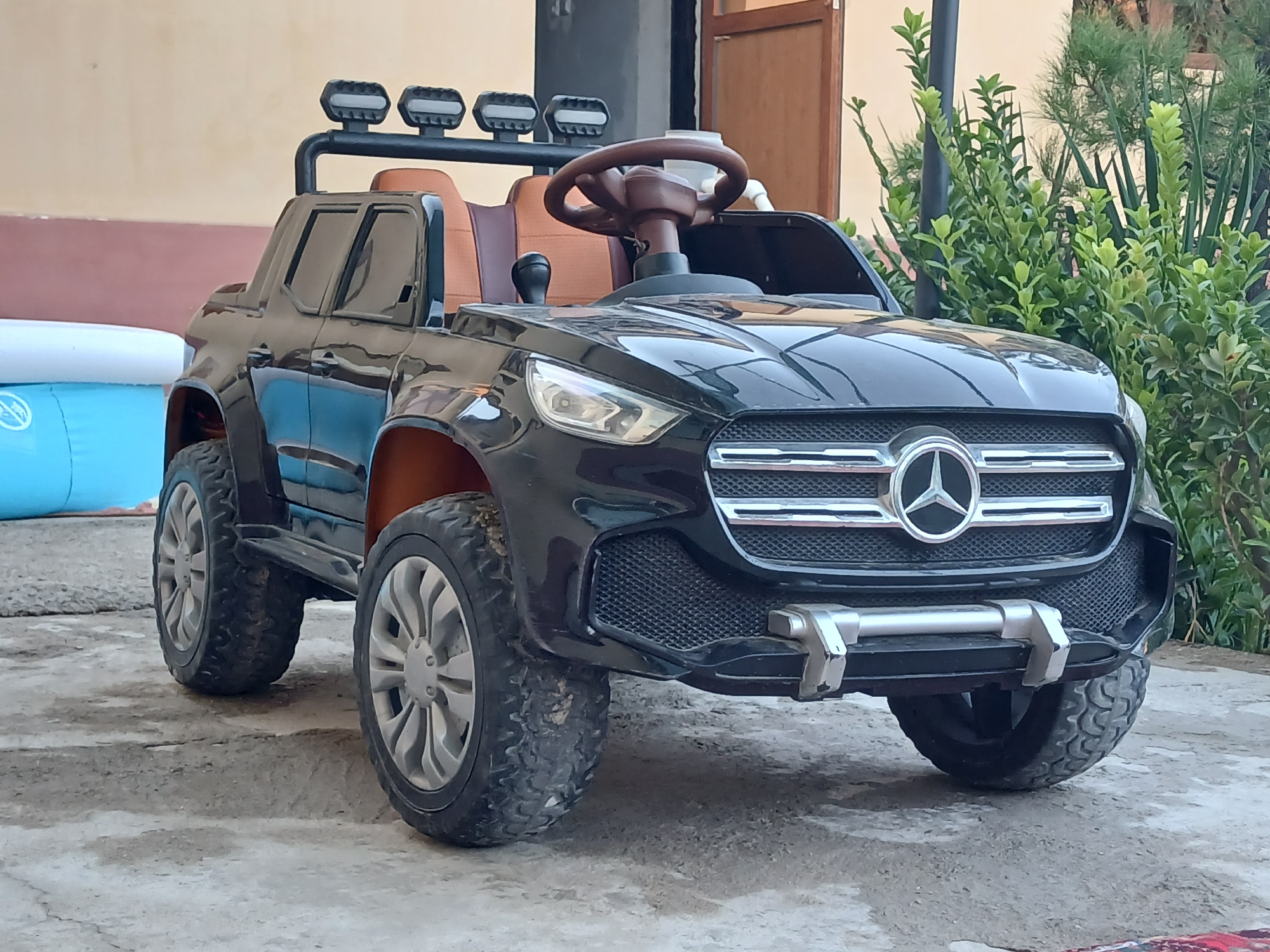 A black Mercedes-Benz ride-on toy car is parked outdoors on a concrete surface in Olovyanka, Uzbekistan. The toy car features details like a black grille with a silver Mercedes-Benz logo, headlights, a brown steering wheel, and orange interior seating. It has large, off-road style tires and a roll bar with four auxiliary lights on top. To the left of the car, a portion of a deflated blue and white inflatable pool is visible. In the background, there is a wall, a wooden door, and lush green bushes and trees. The lighting suggests it is daytime.