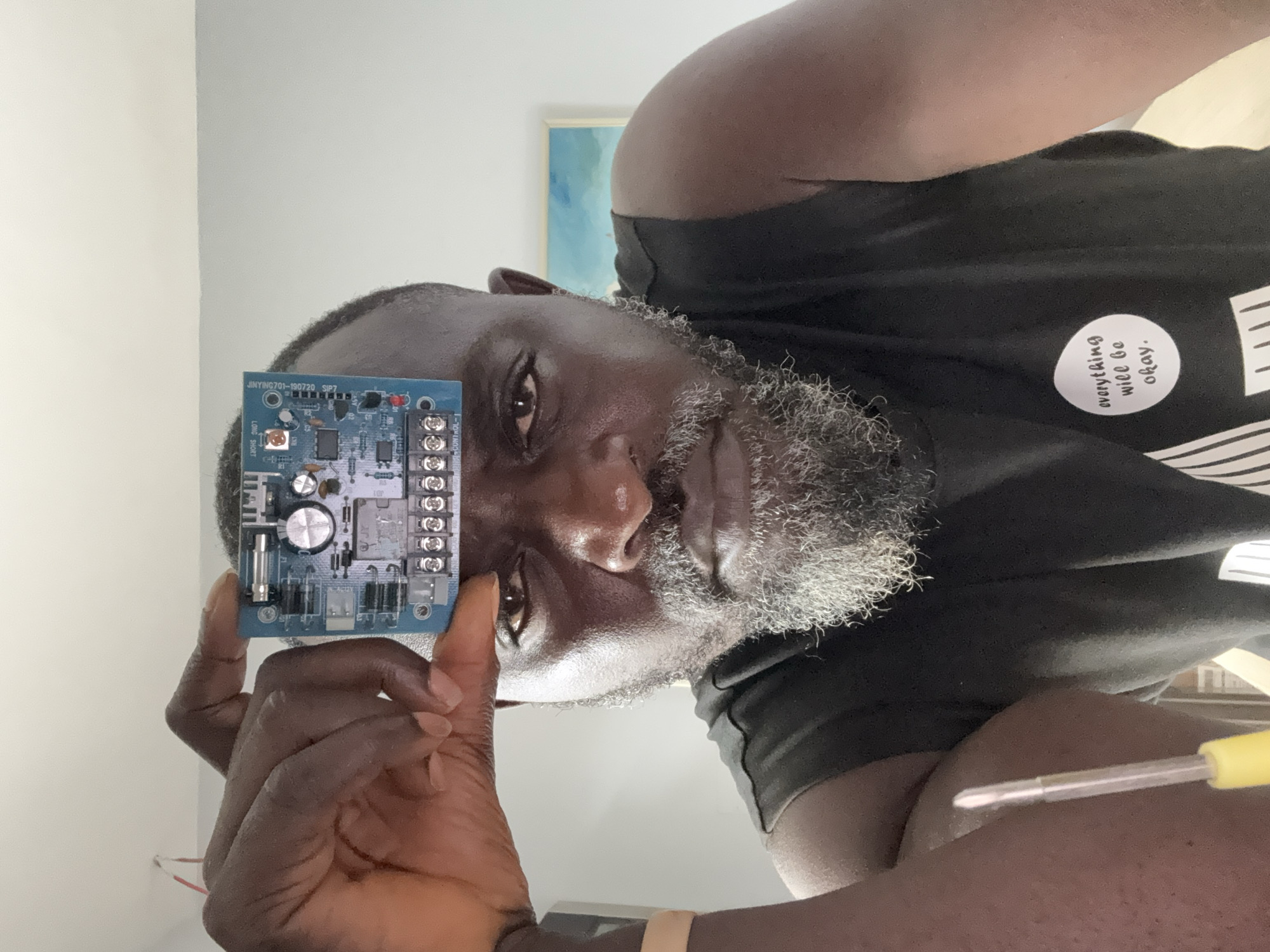 A medium, eye-level shot shows a man holding a blue circuit board between his thumb and index finger. The circuit board is prominently displayed, with components visible. The man is dark-skinned, with a graying beard and a serious expression. He is wearing a black t-shirt with a round white sticker that says "everything will be okay". His right arm is slightly visible, showing him holding a yellow screwdriver. The background includes a white wall and a framed artwork. The picture has been taken from an unusual angle, tilted counter-clockwise, and there is no evidence to indicate that the location is Lagos, Nigeria.