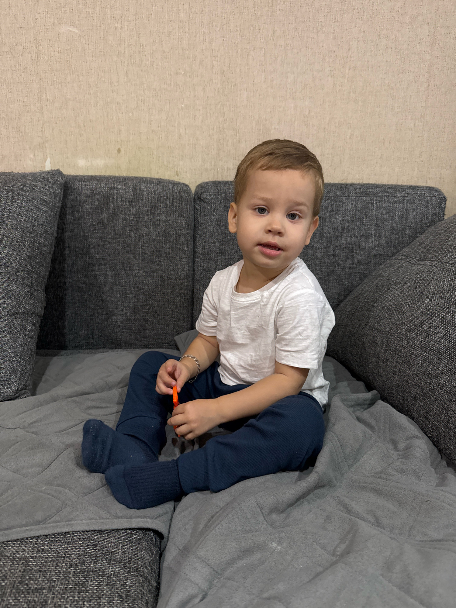 A young Caucasian boy with blond hair and blue eyes is sitting on a gray sectional sofa in a domestic setting. He is wearing a white t-shirt and dark blue sweatpants with dark blue socks. The boy is holding an orange object in his right hand. The sofa is covered with a gray blanket. In the background, a textured beige wall is visible. This scene appears to be located in Rusanovo, Russia, based on the provided information.