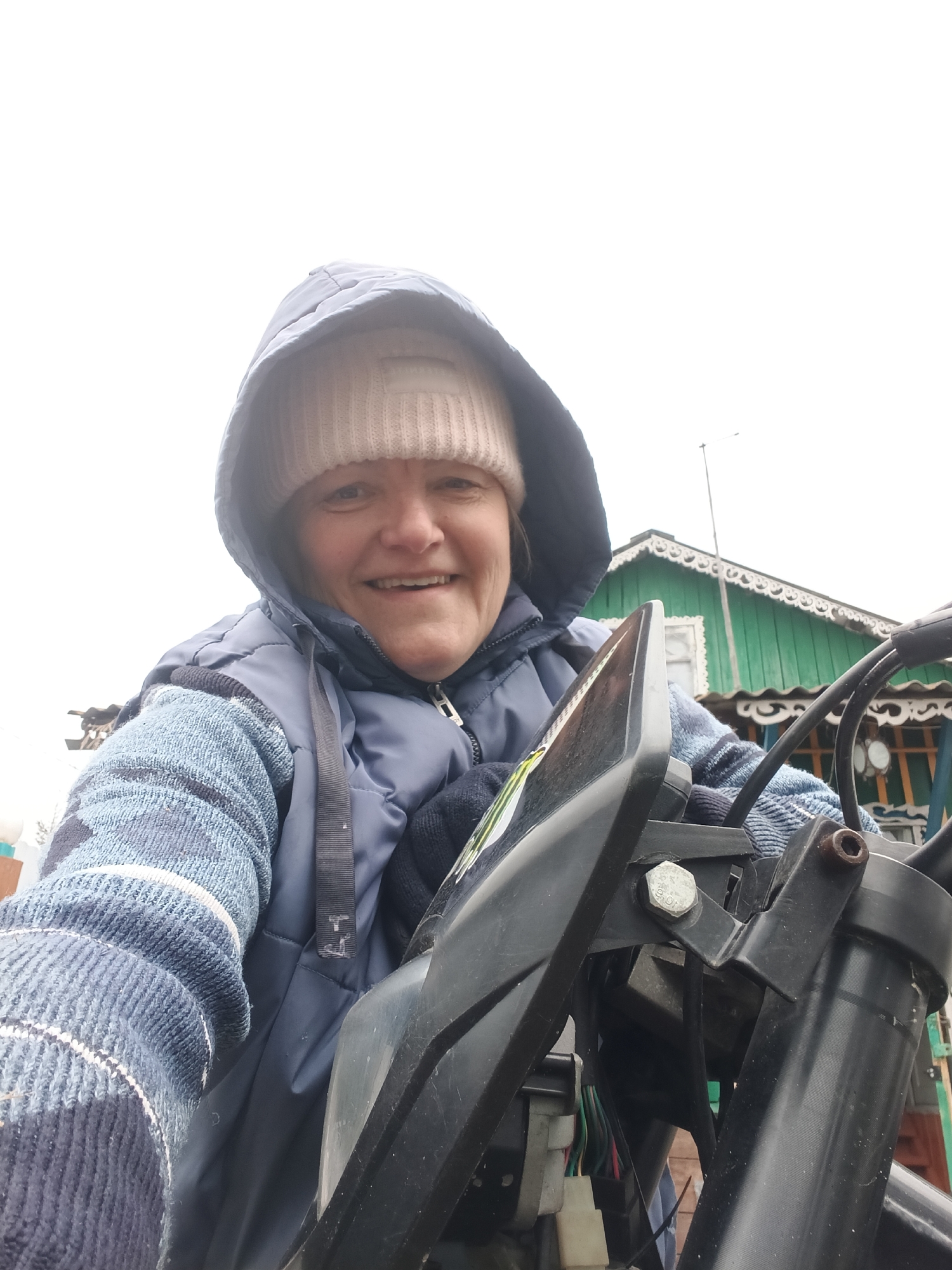 A medium shot depicts a woman, likely in her late 40s or early 50s, seated on a black motorcycle. She wears a light-colored knit cap under the hood of a navy blue puffy vest, and a blue and white patterned sweater beneath. Her expression is cheerful, with a slight smile and her gaze directed toward the camera.
The motorcycle's handlebars and instrument panel are visible in the foreground, providing a point-of-view perspective.
In the background, a green, wooden building with decorative trim and a visible antenna is partially visible.
Based on the metadata, the photograph was taken in Красный Маяк, Russia. The overall impression suggests a casual, outdoor scene.