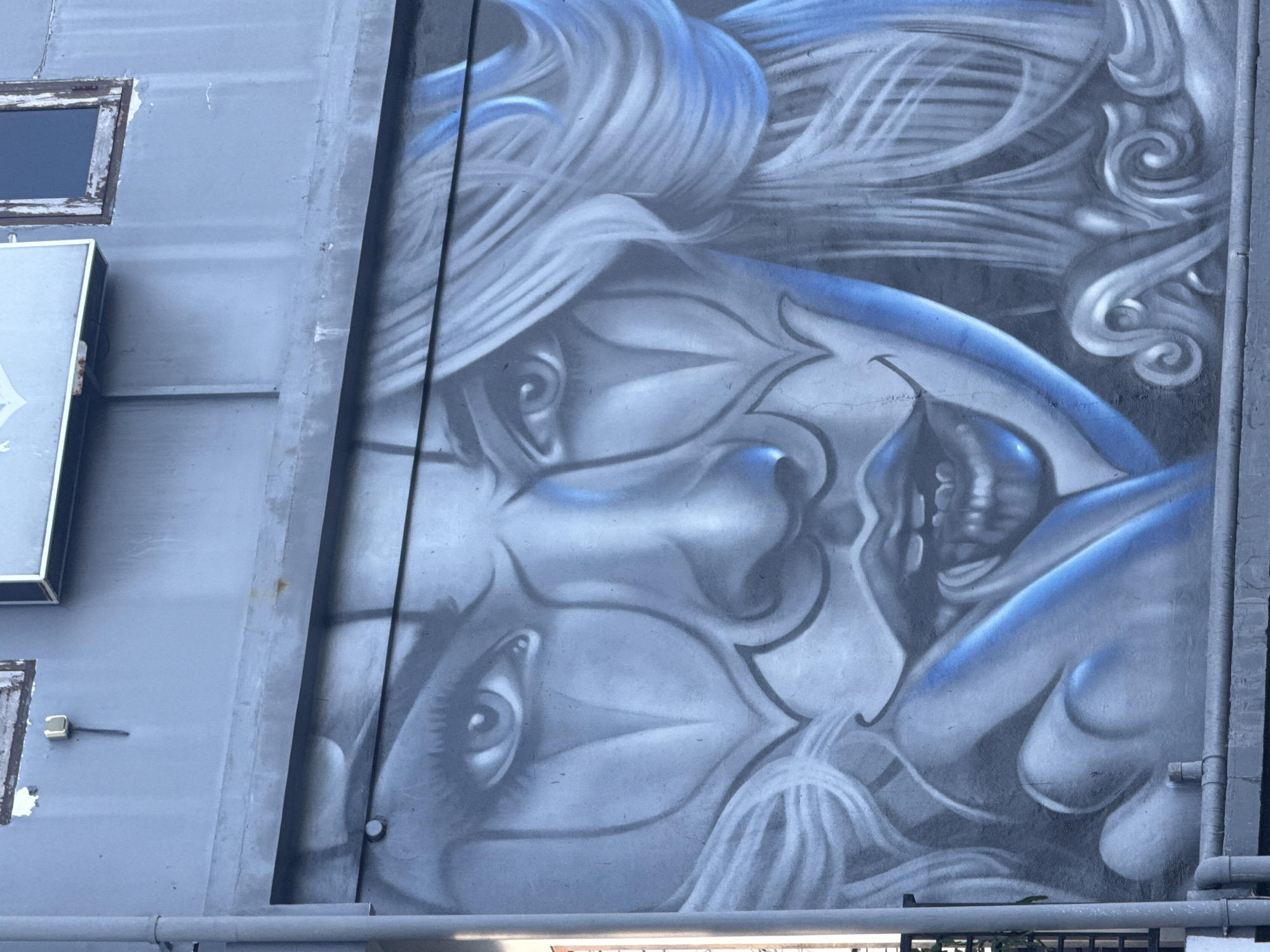 The image depicts a large-scale mural on the exterior wall of a building in Kuta, Indonesia. The mural features a stylized face with blue and white coloring, intricate details around the eyes and nose, and swirling patterns in the hair. The building is constructed of gray metal panels, with pipes running vertically along the right side of the mural. A small window and a sign are visible on the left side of the building. A pipe runs horizontally in the foreground.