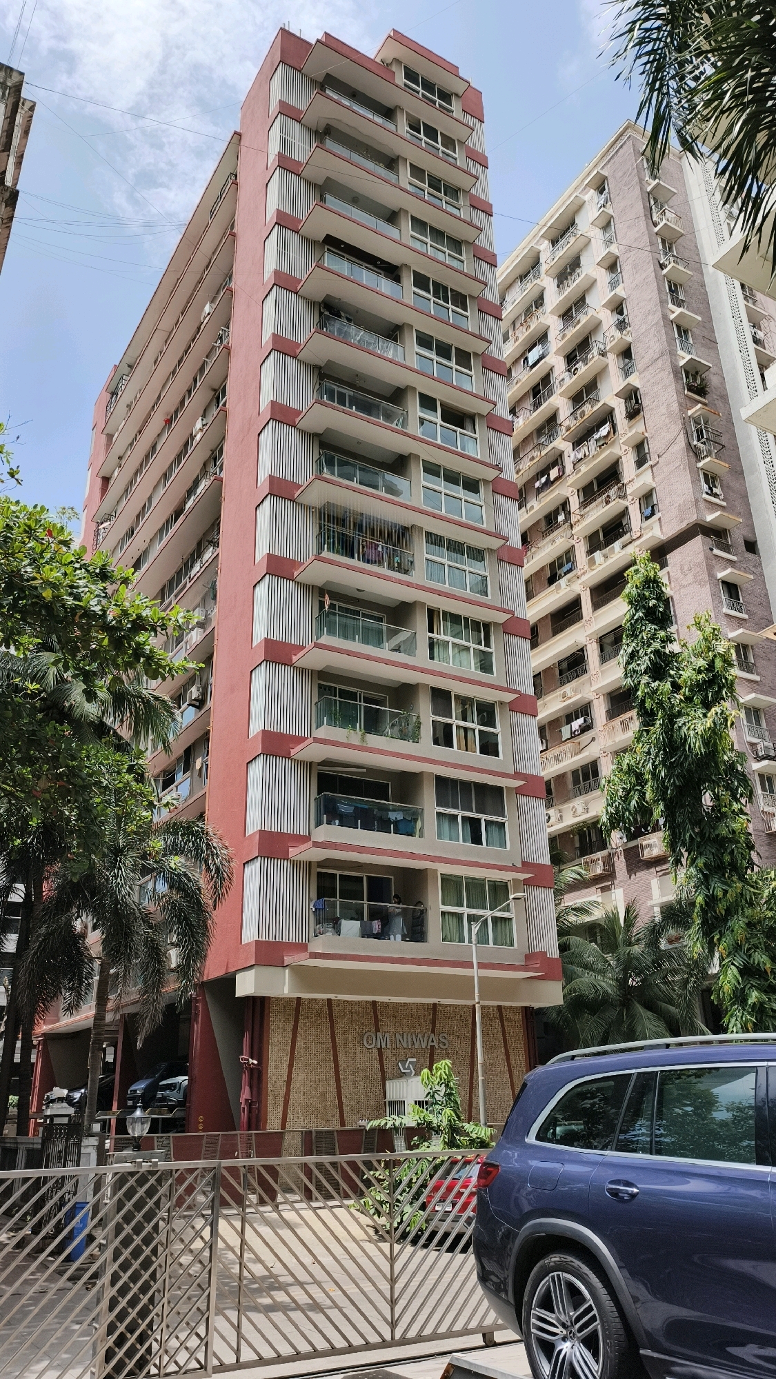 An exterior, medium shot captures the "Om Niwas" apartment building in Mumbai, India, under a cloudy sky. The building, painted in a salmon pink and maroon color scheme, features a rhythmic pattern of balconies with slatted shades and glass railings, interspersed with windows. Adjacent to it, another taller, light-pink building with numerous balconies and laundry hanging out is visible. Palm trees and other lush green foliage surround the base of the buildings, with a dark blue SUV parked in the foreground, partially obscuring a metal gate. The signage "OM NIWAS" is clearly visible on the lower facade of the salmon-pink building.