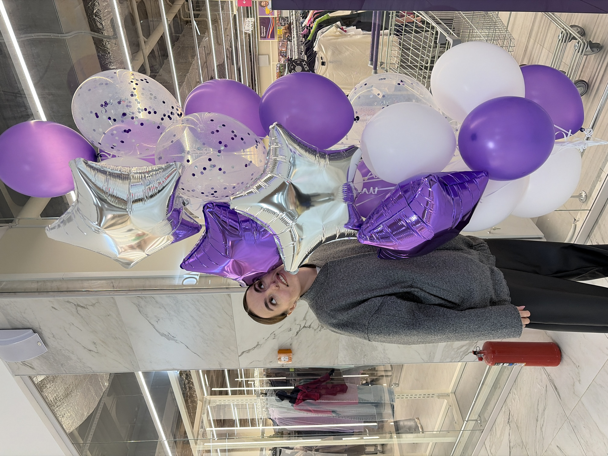 A person is standing in what appears to be a retail store in Izhevsk, Russia. They are holding a large bouquet of balloons in white, purple, and silver, including star-shaped foil balloons and clear balloons with confetti. The balloons obscure part of the person's face. The individual is wearing a gray sweater and black pants. The store environment has clothing racks with merchandise visible behind a clear display case, a fire extinguisher on the floor, and overhead lighting fixtures. A shopping cart is partially visible in the background.